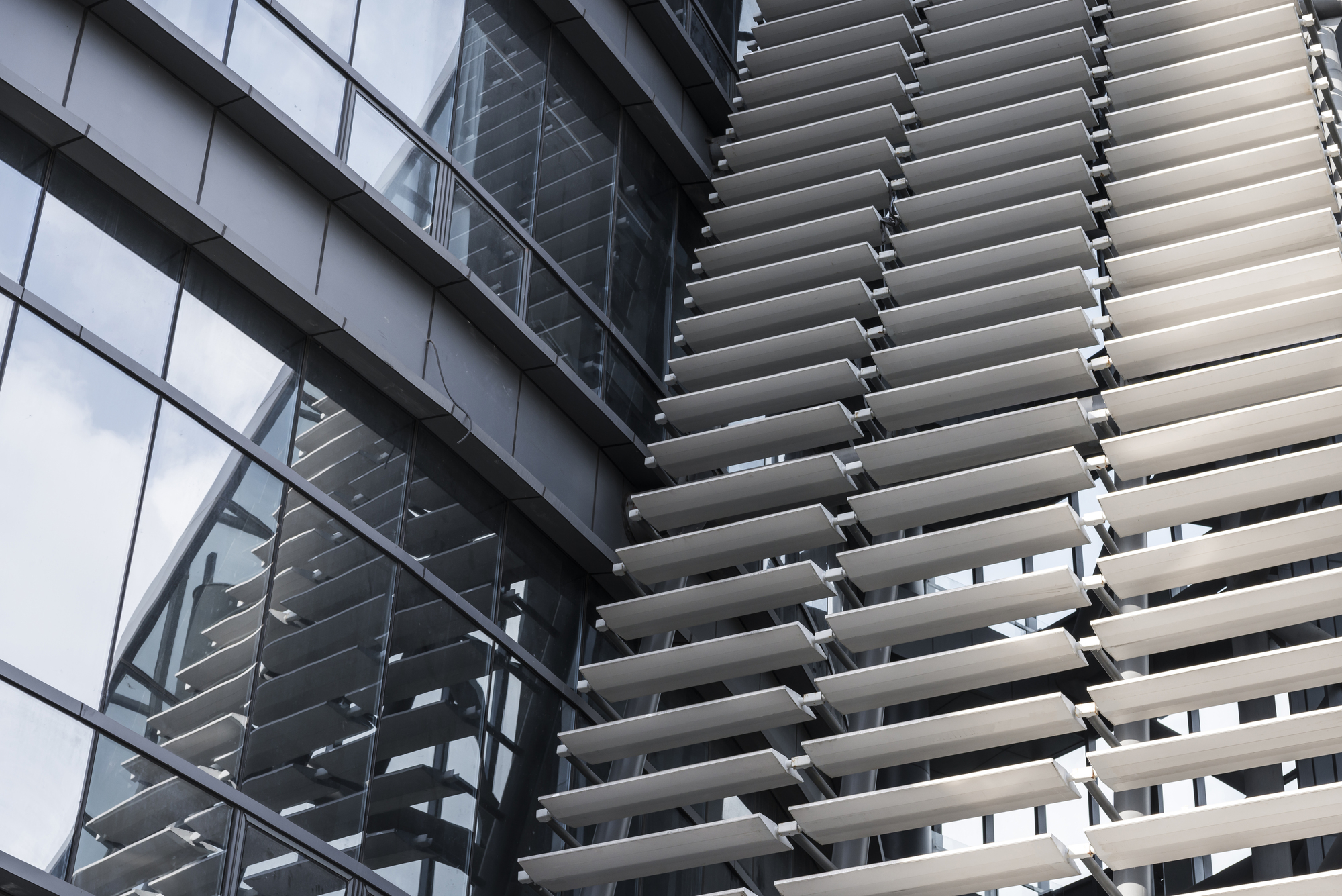 Angled aluminum louvers stacked beside a glass curtain wall that reflects the louvers and a cloudy sky