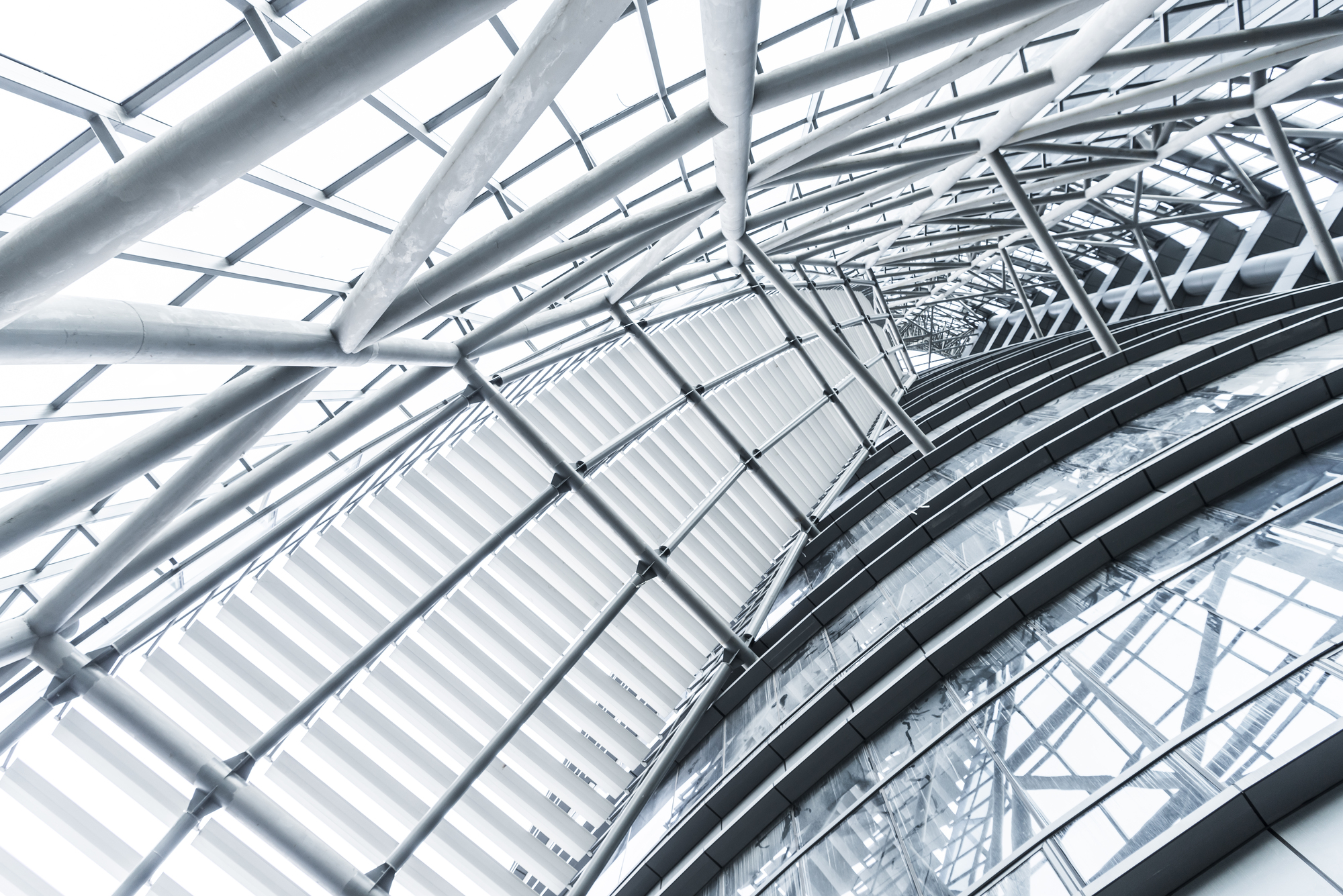 Upward view of tubular steel trusses and curved glazing forming a vaulted atrium roof in cold white light