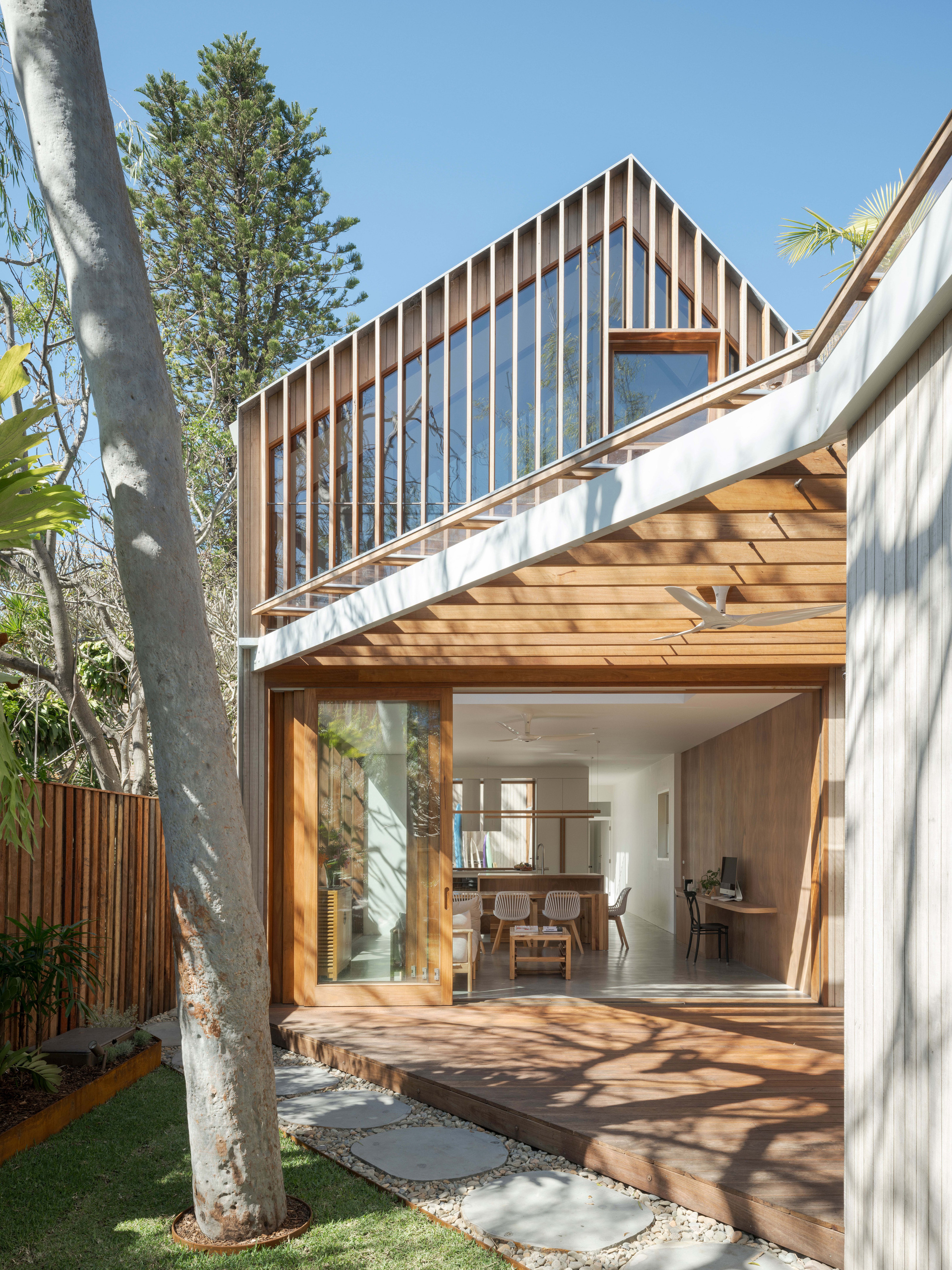 Rear extension with a gabled upper level of timber fins and glazing, open sliding doors to a deck, and a pale tree trunk in front