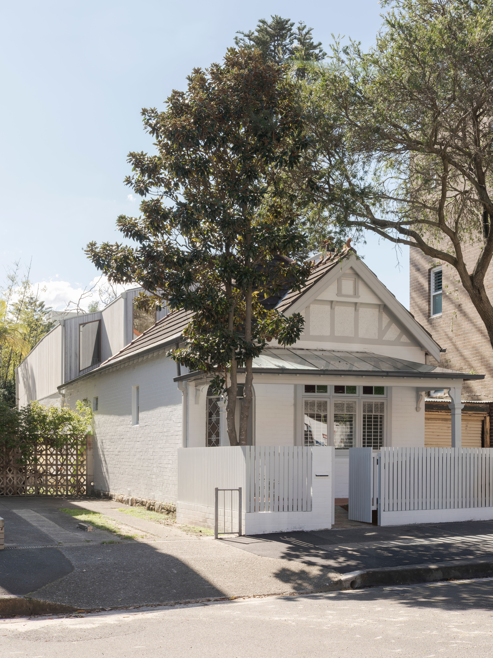 Street view of a white painted brick cottage with a picket fence and a timber-clad upper addition behind a magnolia tree