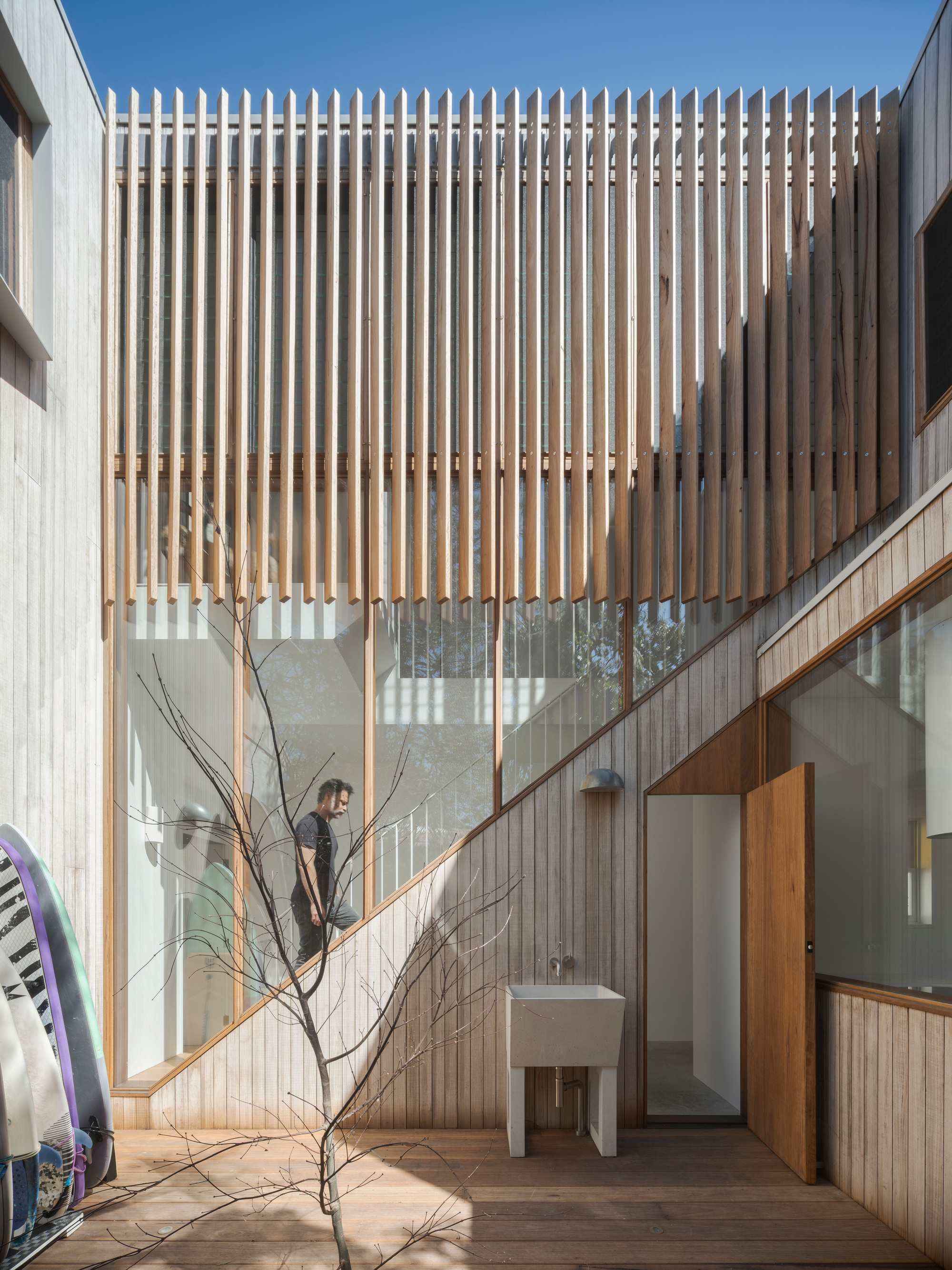 Timber-lined courtyard with a slatted screen above glazing, an outdoor concrete sink and a man climbing the stair beside surfboards