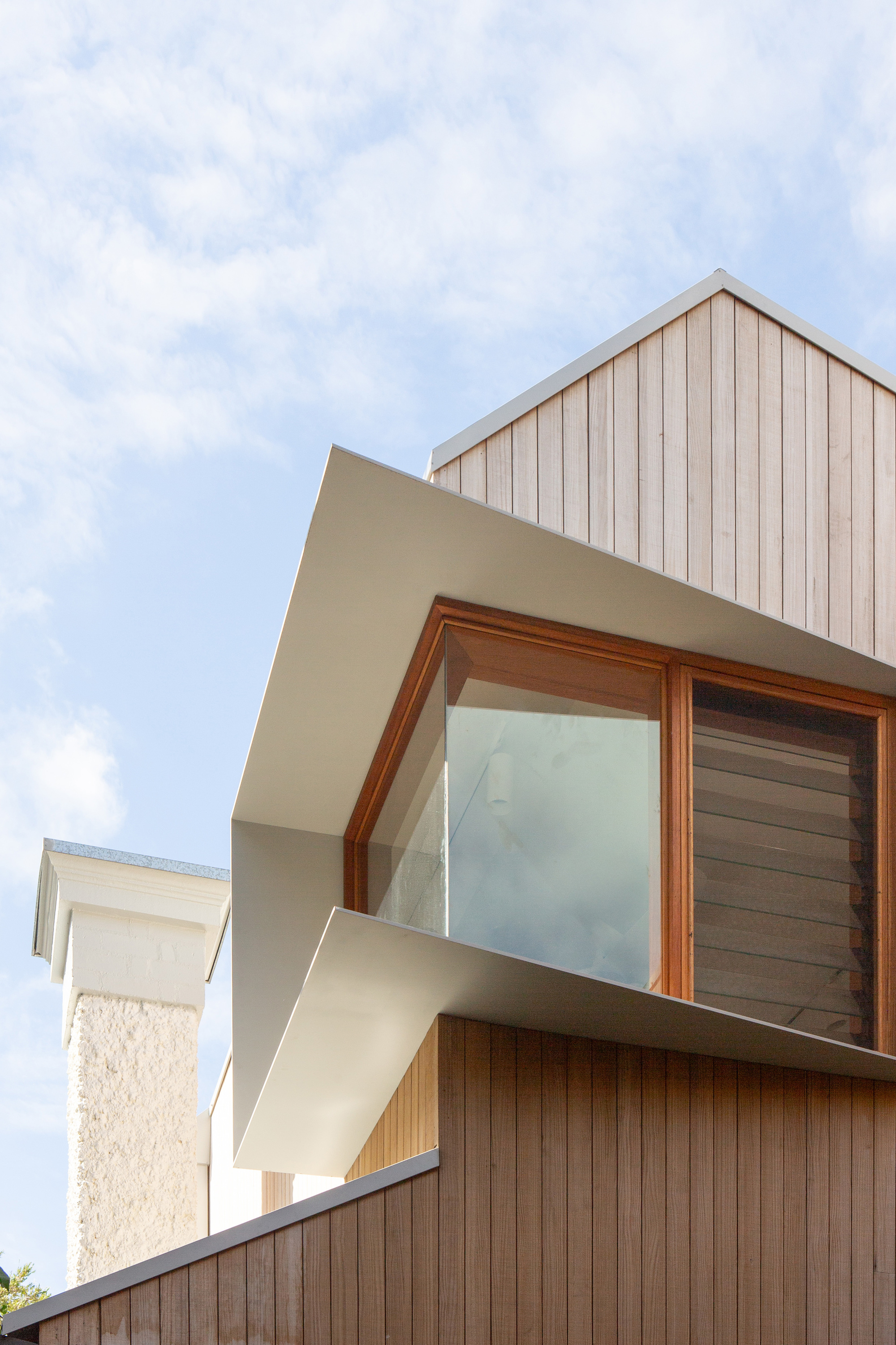 Detail of the pale metal-framed corner window and vertical timber cladding beside a neighbouring chimney under a cloudy sky