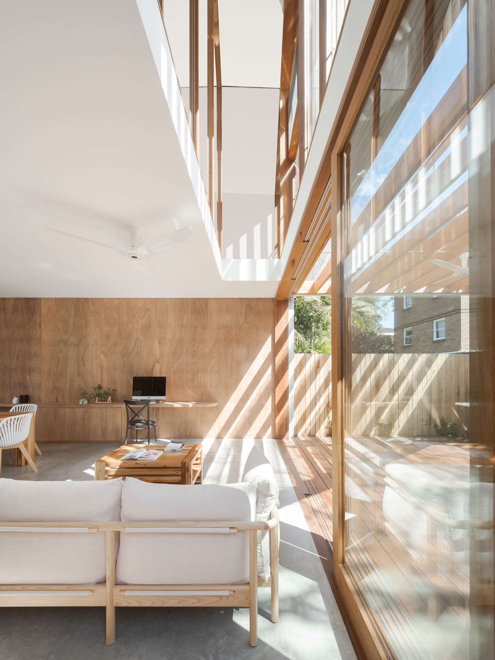Living area with plywood wall, low timber sofa and double-height void, with sunlight casting striped shadows across the concrete floor from sliding glass doors