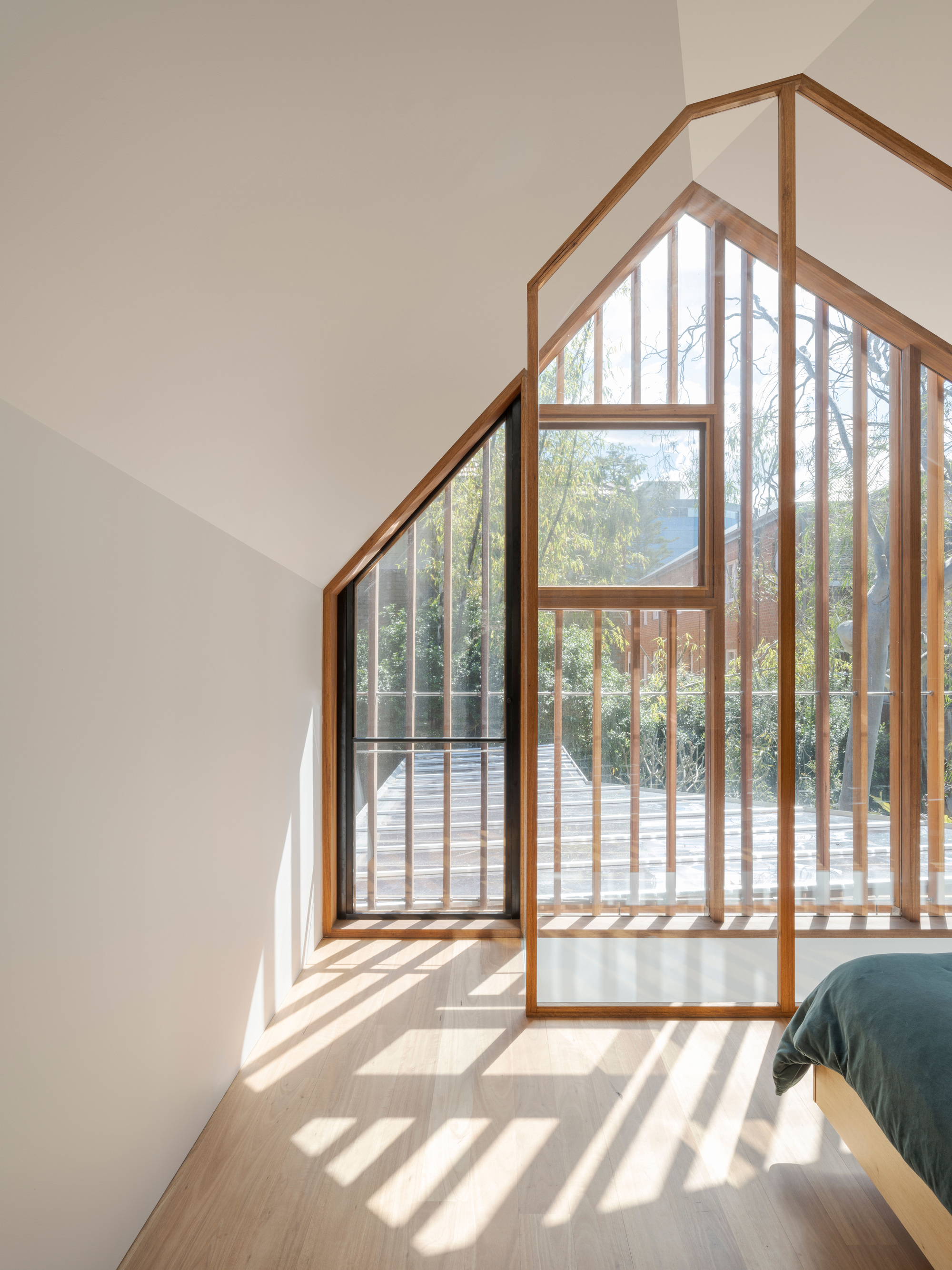 Upstairs bedroom with a gable-shaped timber-framed window and vertical fins, sunlight striping the oak floor beside the corner of the bed