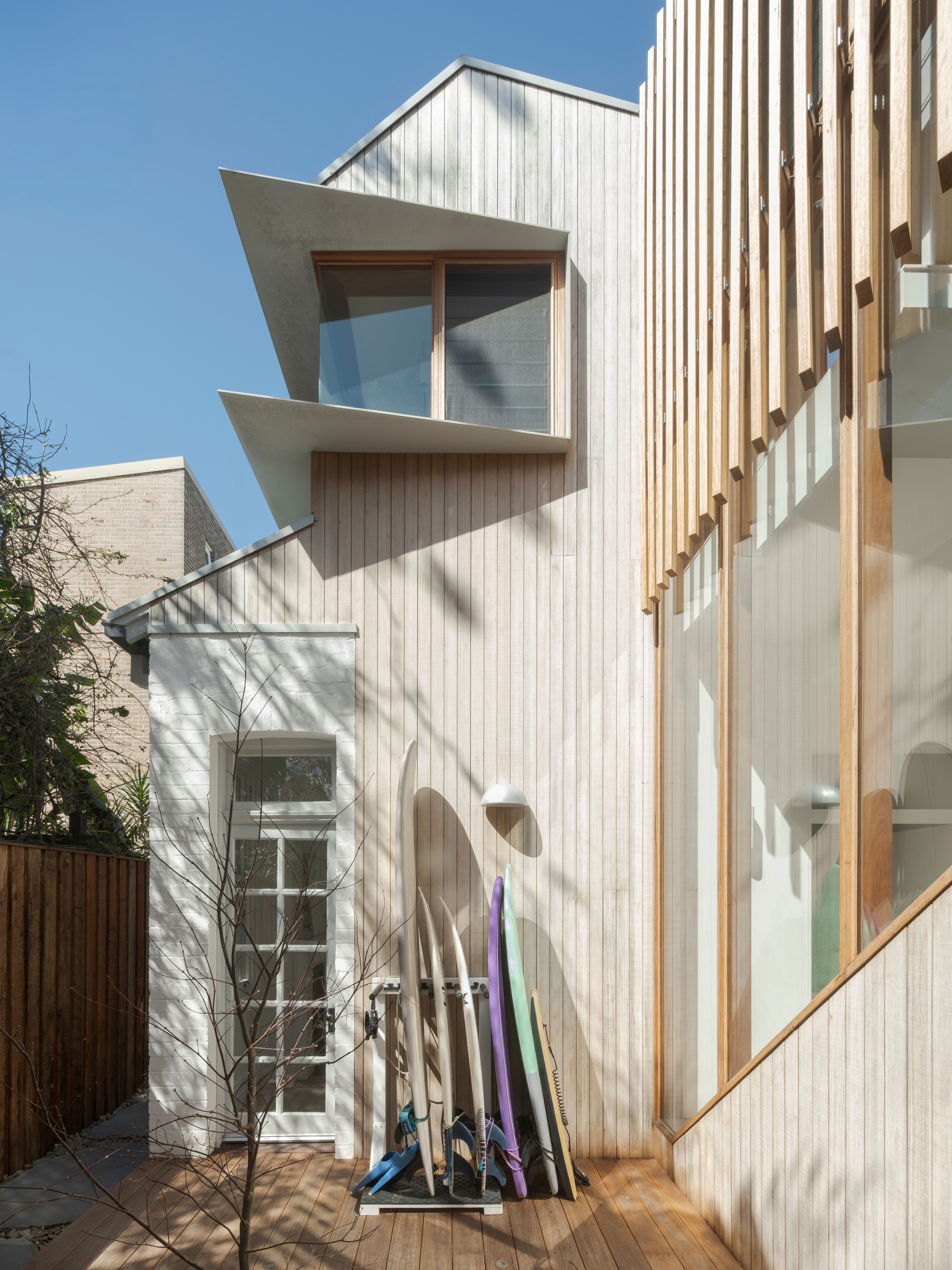 Angular concrete-edged bay window above a white painted doorway, with surfboards leaning on the deck in hard midday sun