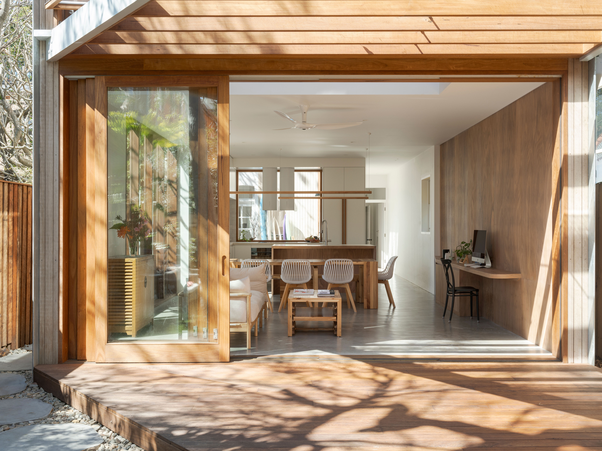 Sliding timber-framed glass doors opened from the deck to a kitchen and dining space, with tree shadows on the boards