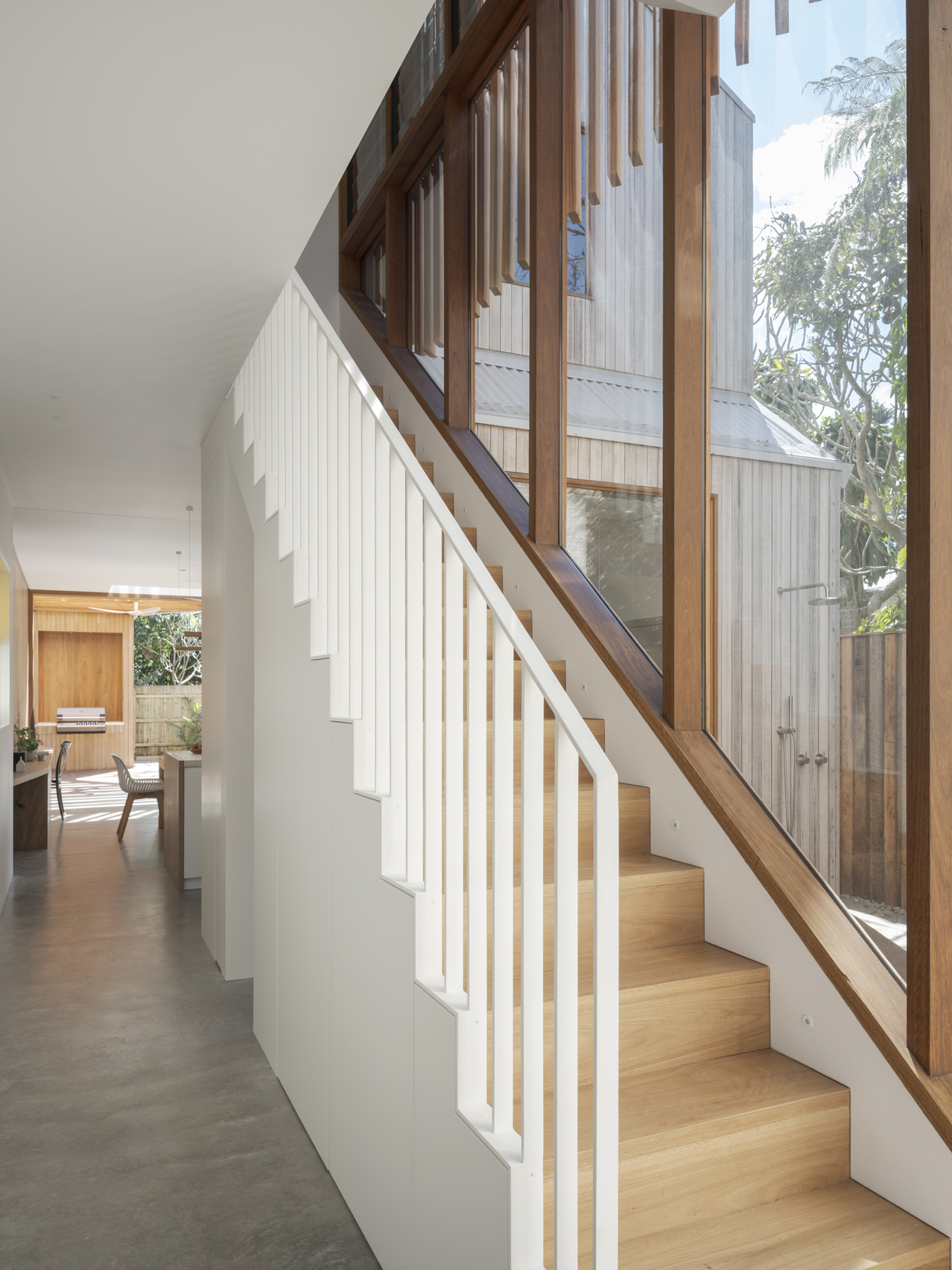 White balustraded staircase with timber treads beside a long window, and a concrete-floored hallway running back toward the kitchen