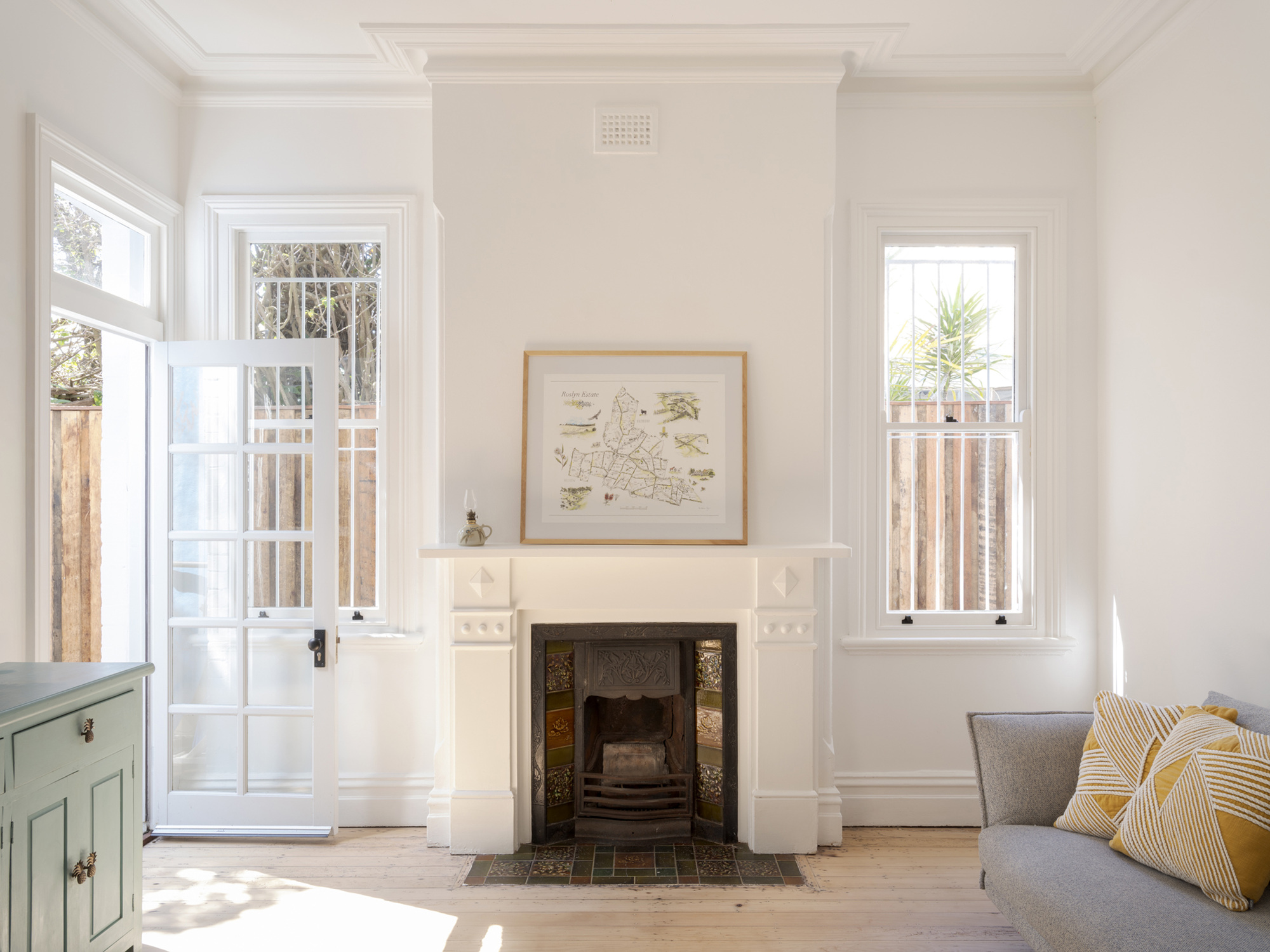 Living room with a white mantelpiece and cast iron fireplace, a framed map above, and French doors open to a sunlit fence