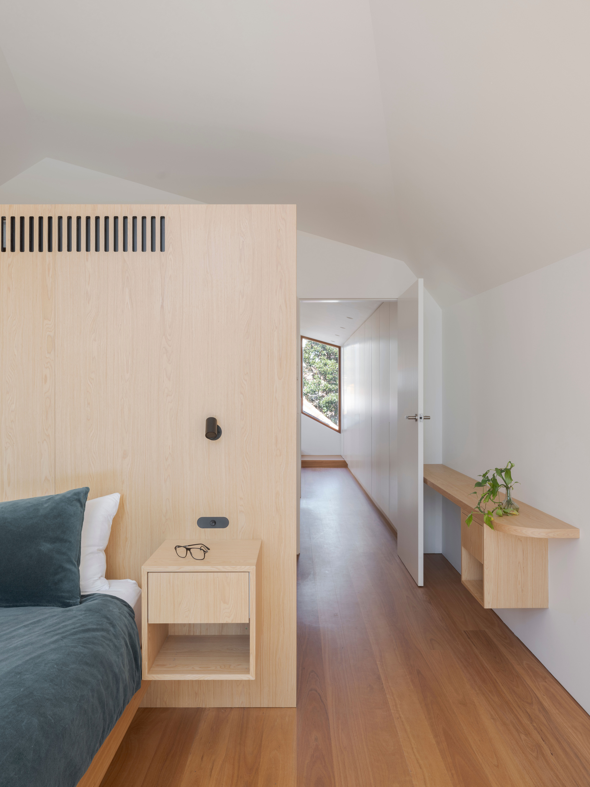 Bedroom with a timber headboard partition, floating bedside table and a corridor leading to a angled window in afternoon daylight
