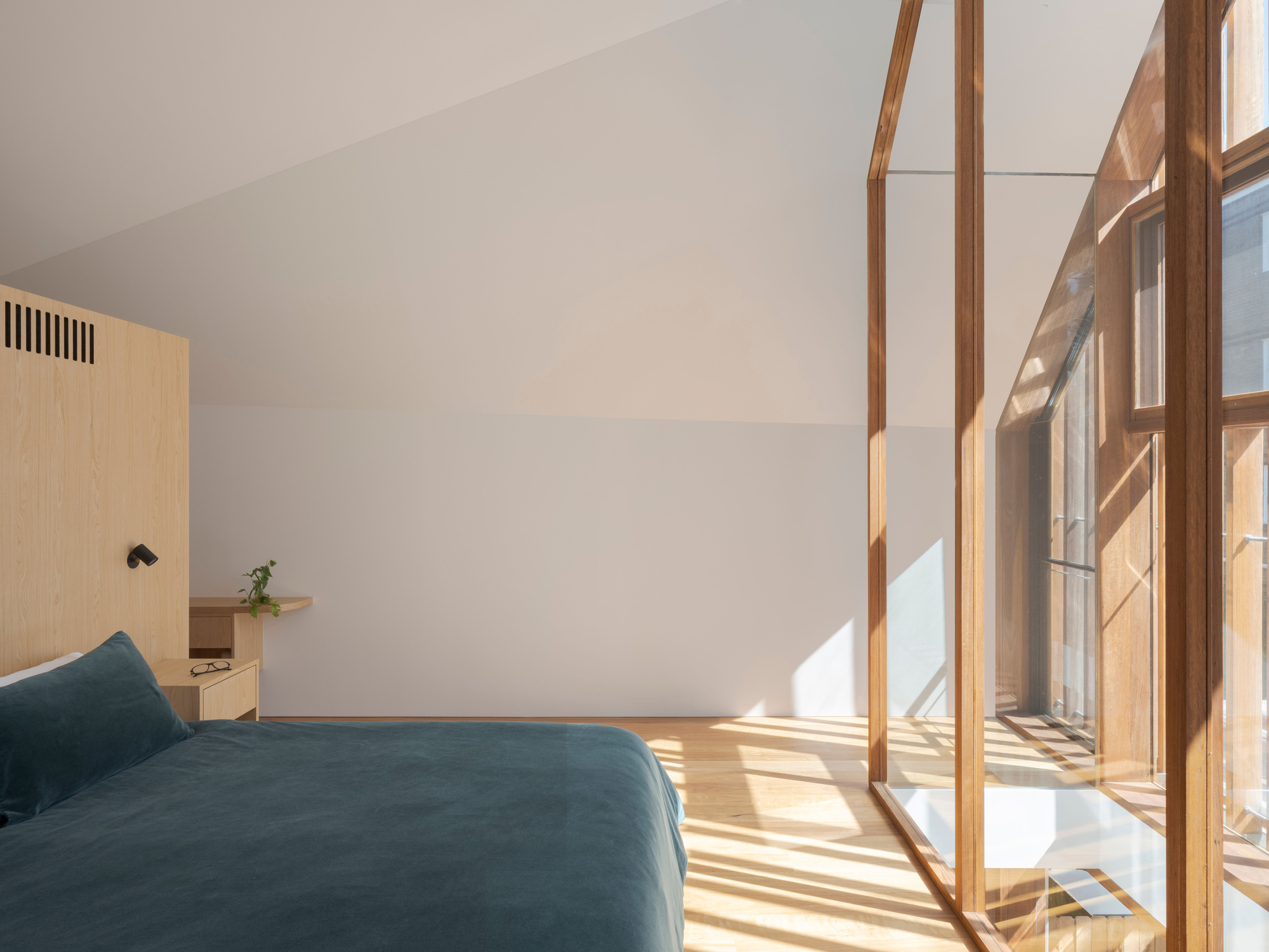 Bedroom beside a timber-framed glass wall, with sunlight casting long stripes across the oak floor and a teal bed cover