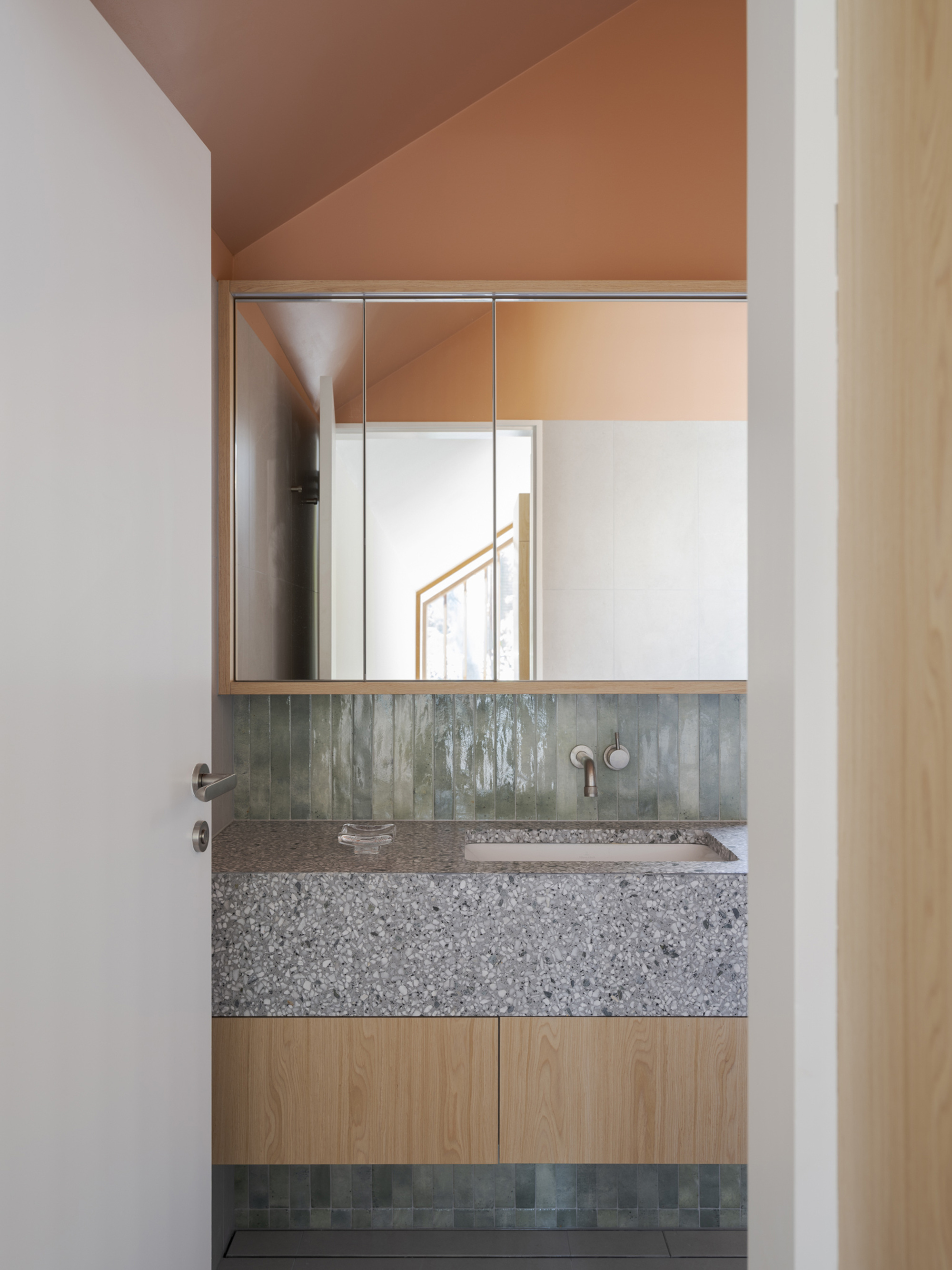 Bathroom vanity with terrazzo-style stone top, green glazed tile splashback and mirrored cabinet under a peach-coloured sloped ceiling