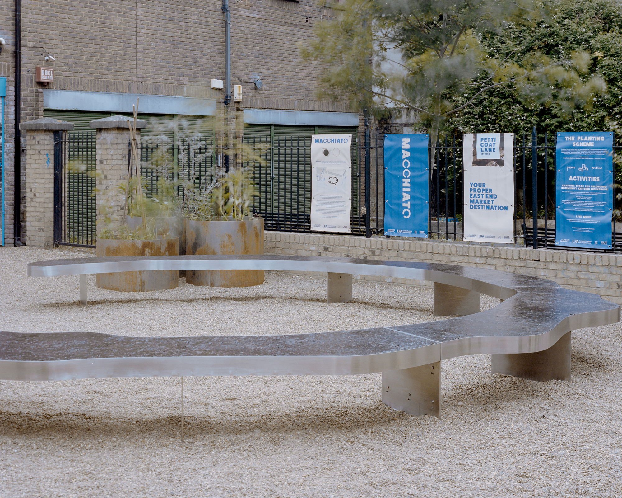 Curving stainless steel bench with a stone top in a gravel court, rusted planters and hanging banners on the fence behind