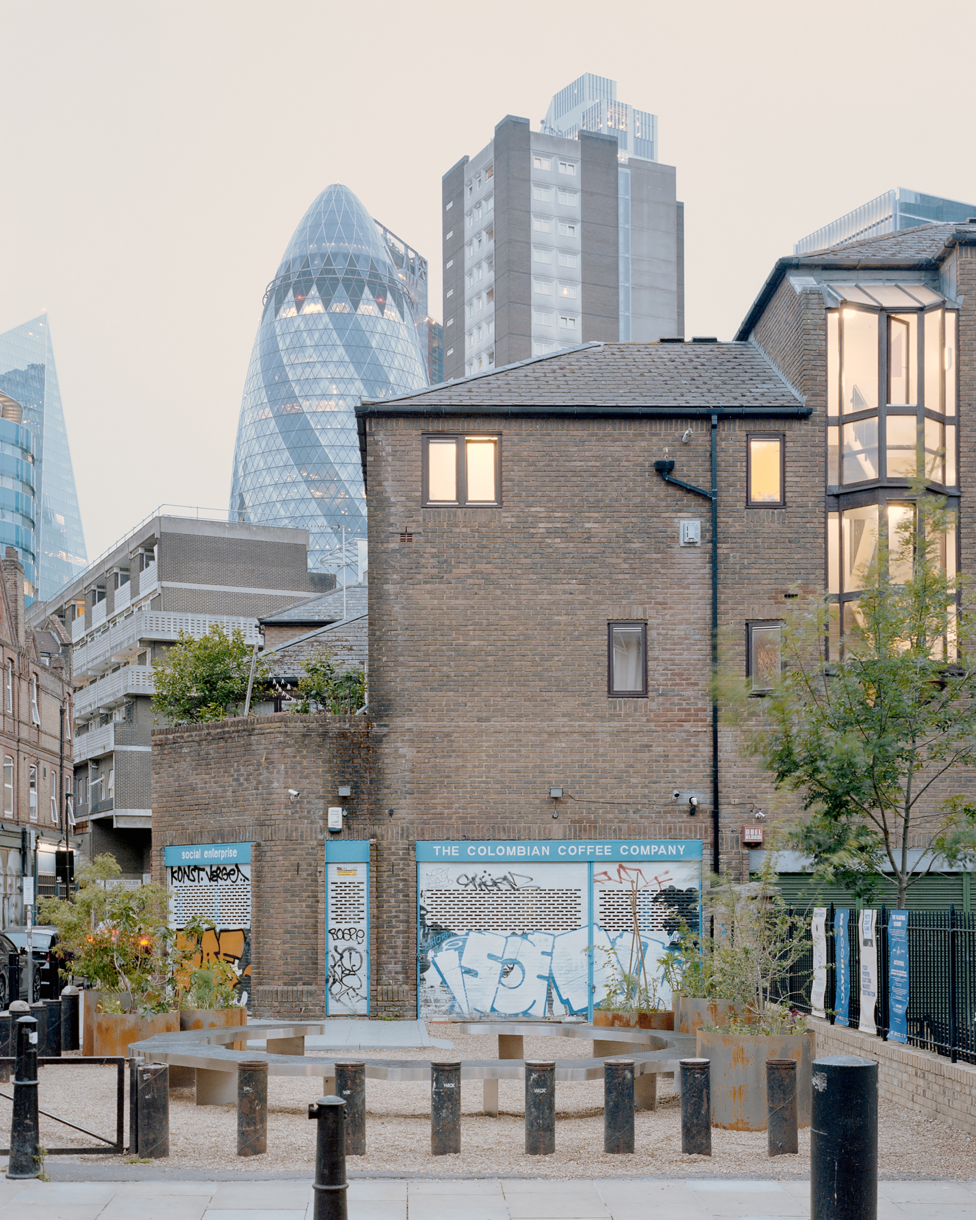 Brick backs of buildings and a graffitied shopfront beyond a gravel court, a glass tower rising behind at dusk