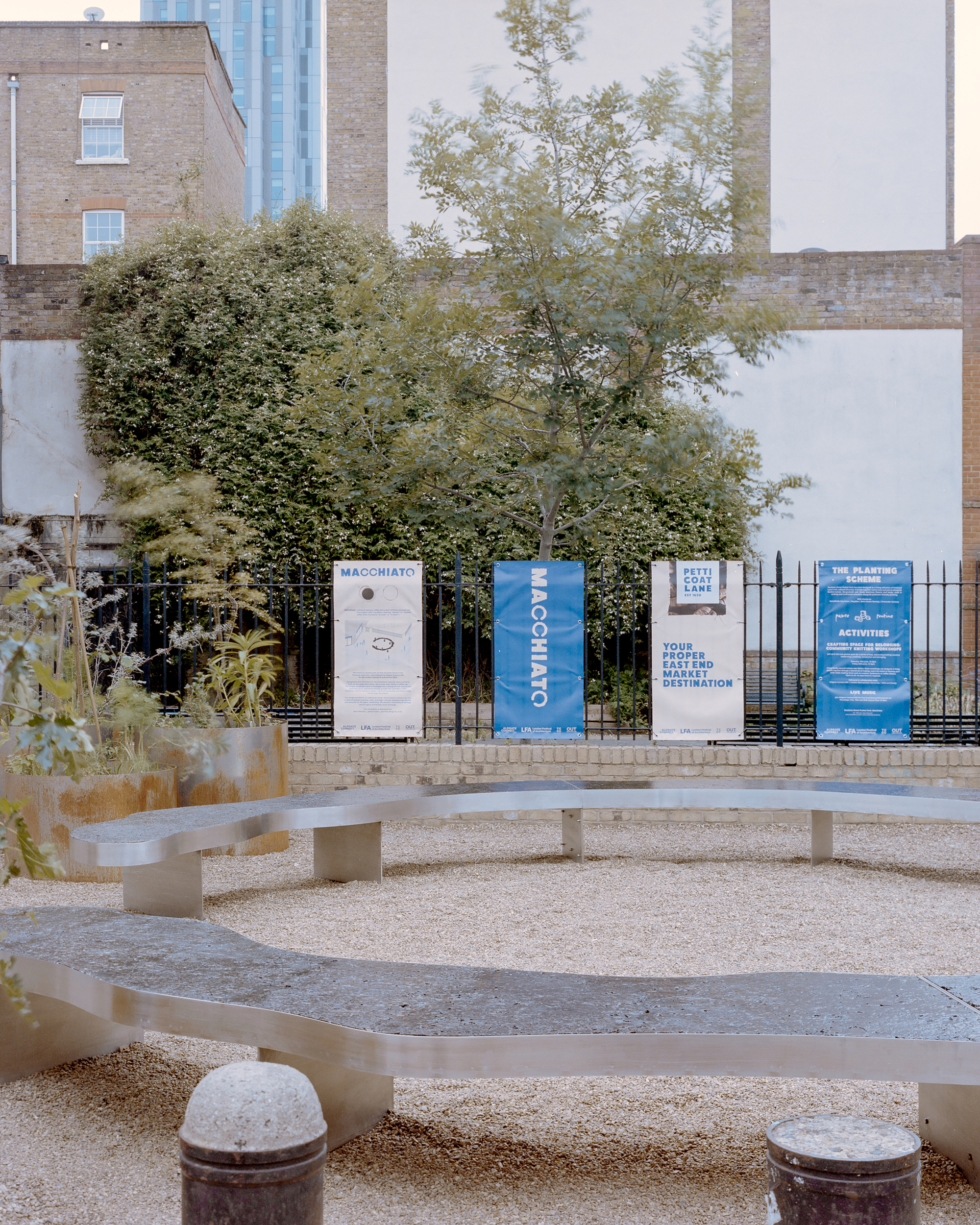 Wavy steel benches on gravel in front of a railing hung with banners, a tree and white party wall behind