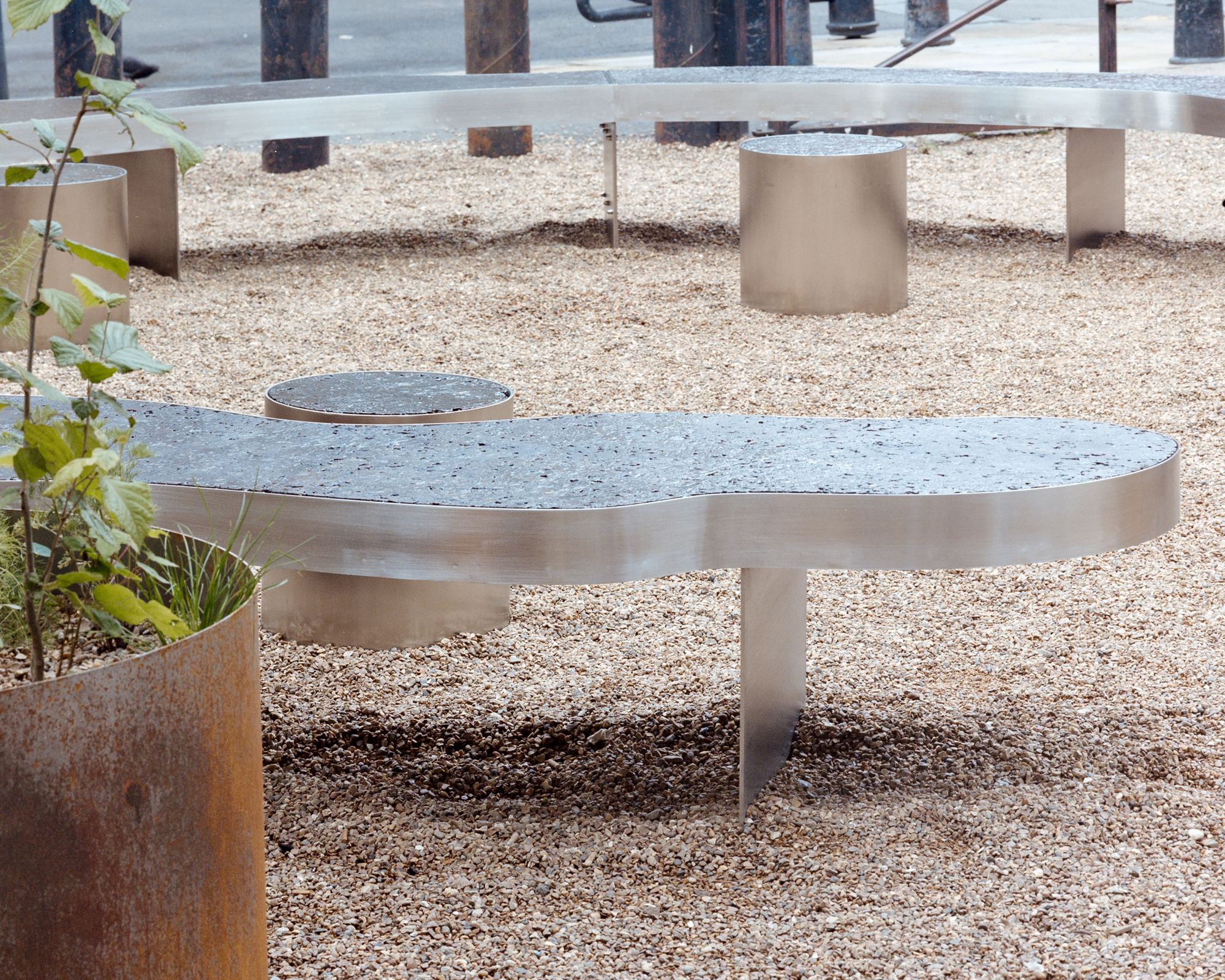 Low steel bench with a stone top and cylindrical stools on pale gravel beside a rusted planter holding a young tree