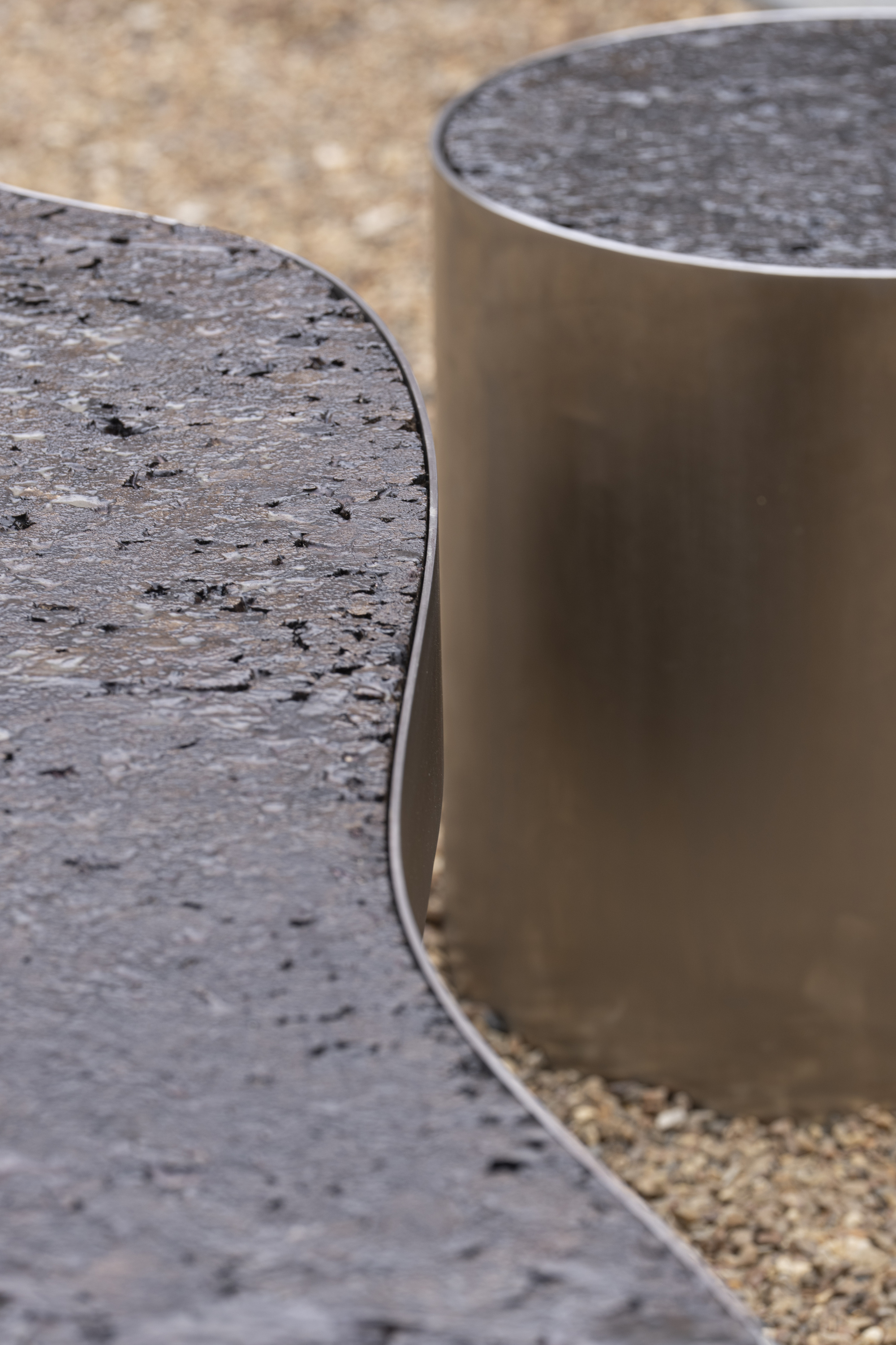 Close-up of the textured stone bench top, its curved steel edge and a brushed steel cylindrical stool above gravel
