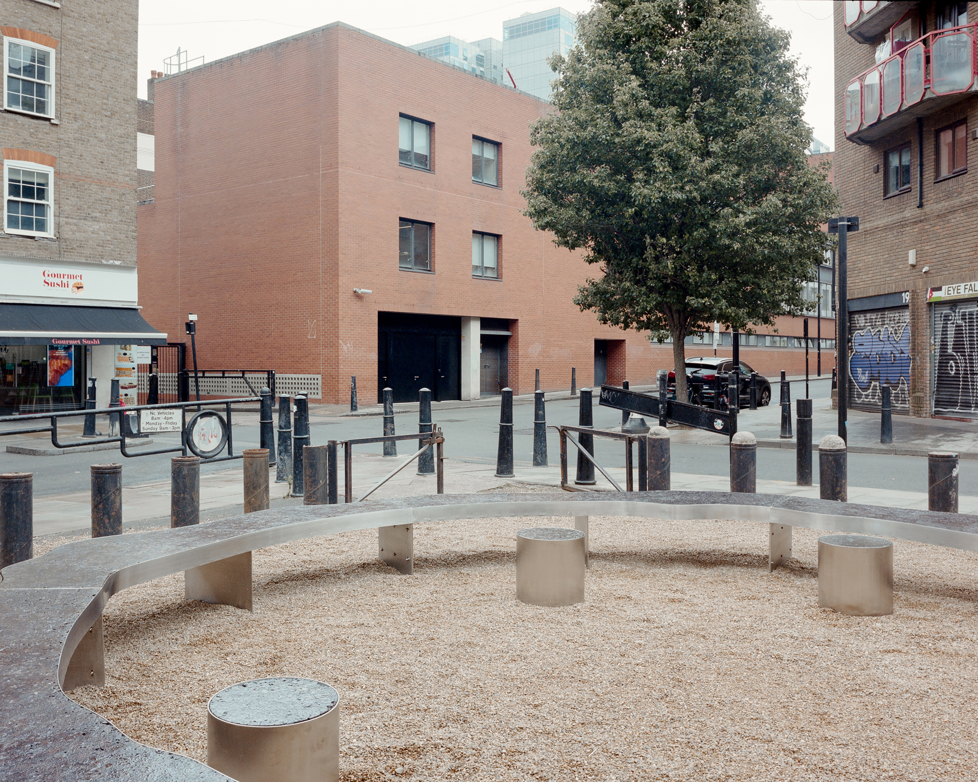 Long curved bench and steel stools on wood chip ground, with bollards, a red brick building and a street tree beyond