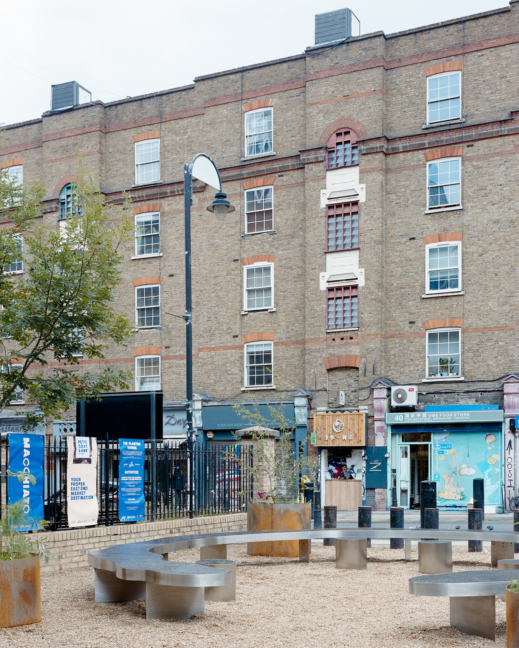 Curved steel benches and rust-colored planters on a gravel square in front of a yellow brick housing block and shopfronts, under overcast sky