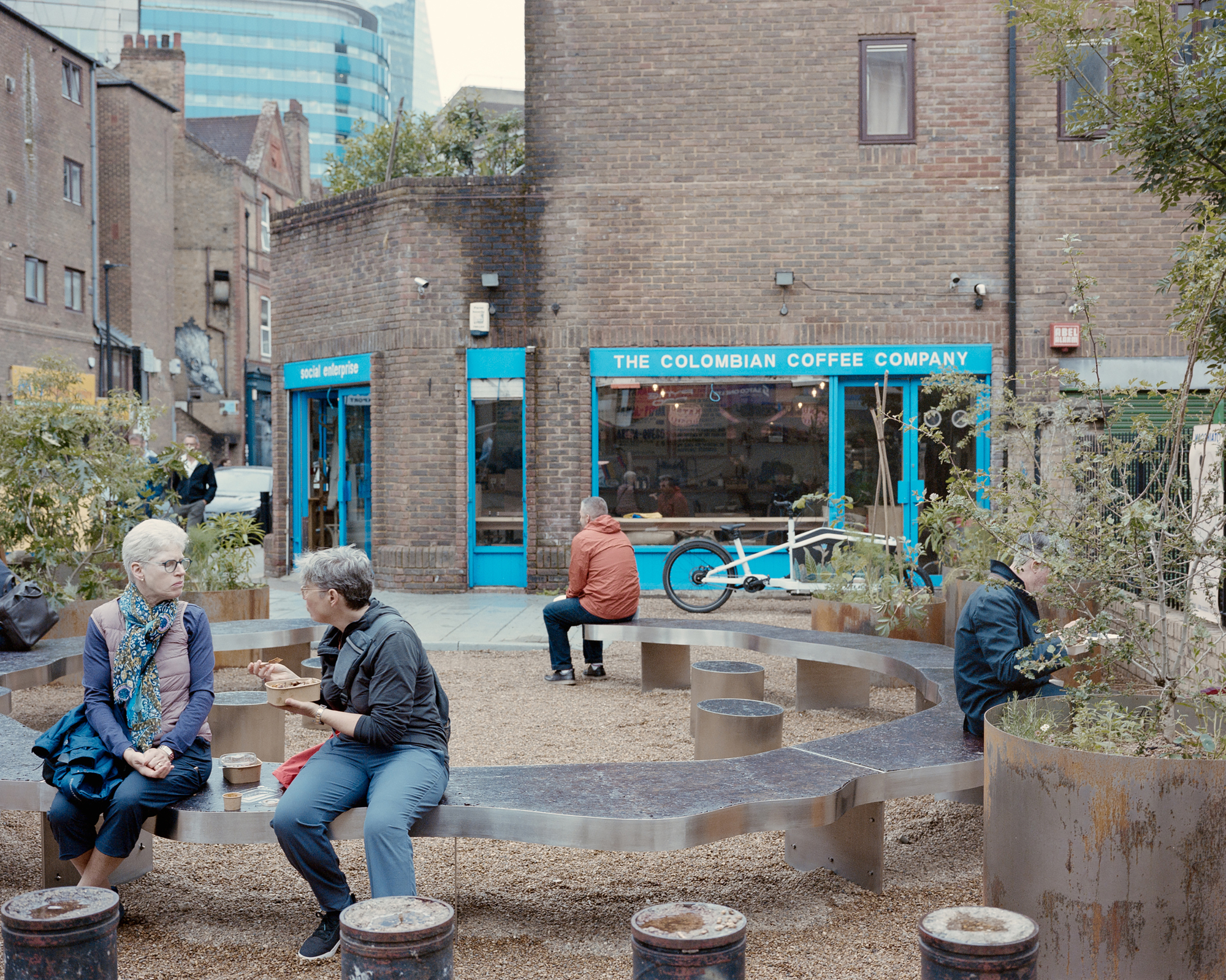 Two women talking on a wavy steel bench in front of a blue-framed coffee shop, with a man seated beyond