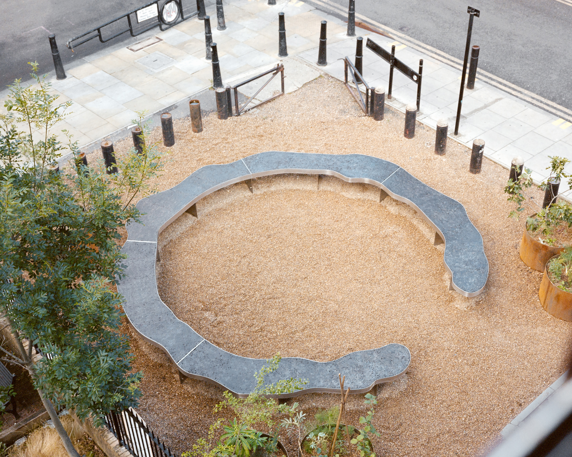 High view of the open ring of undulating steel bench set in gravel, edged by bollards and a paved pavement