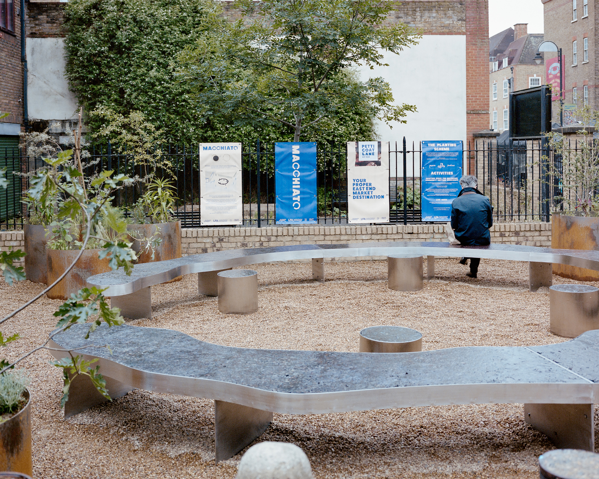 Man seated on the curving steel bench facing an iron fence hung with event banners, with round stools and planters nearby