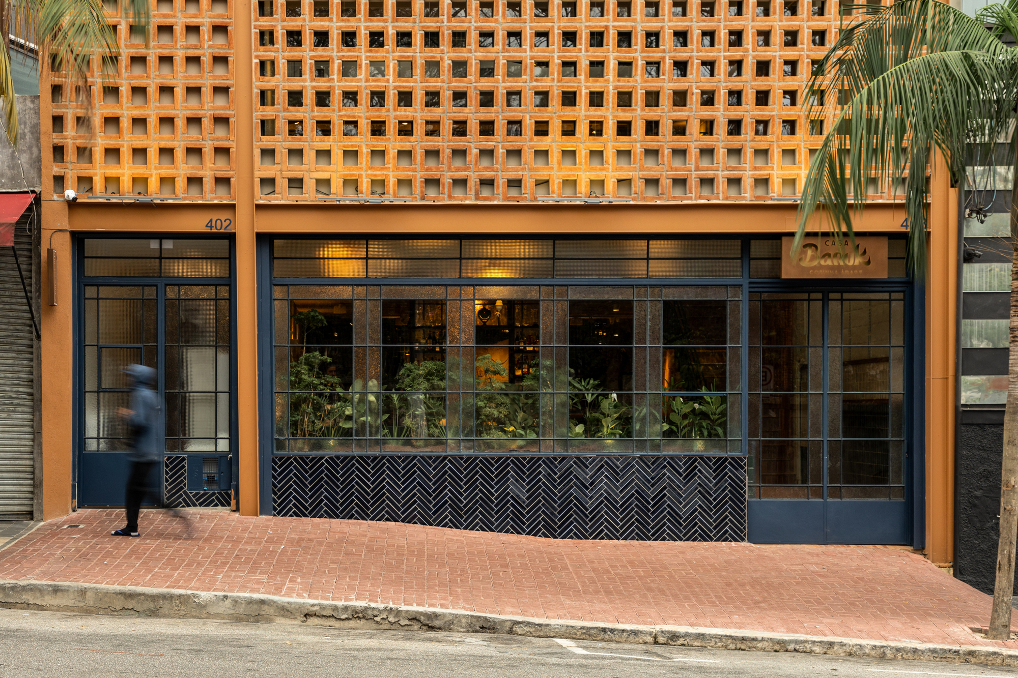 Street facade with an orange perforated block screen above blue steel-framed glazing and a herringbone tile base, a pedestrian passing on the brick sidewalk