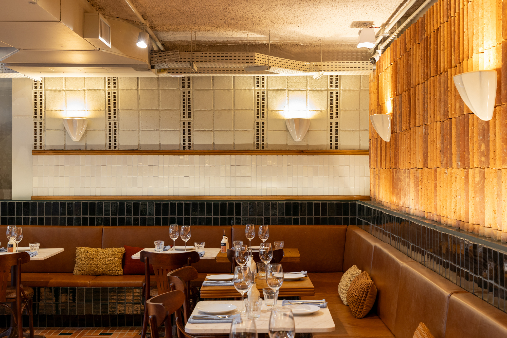 Dining corner with leather banquette, dark green tile dado and a wall of vertical terracotta bricks glowing under warm sconce light