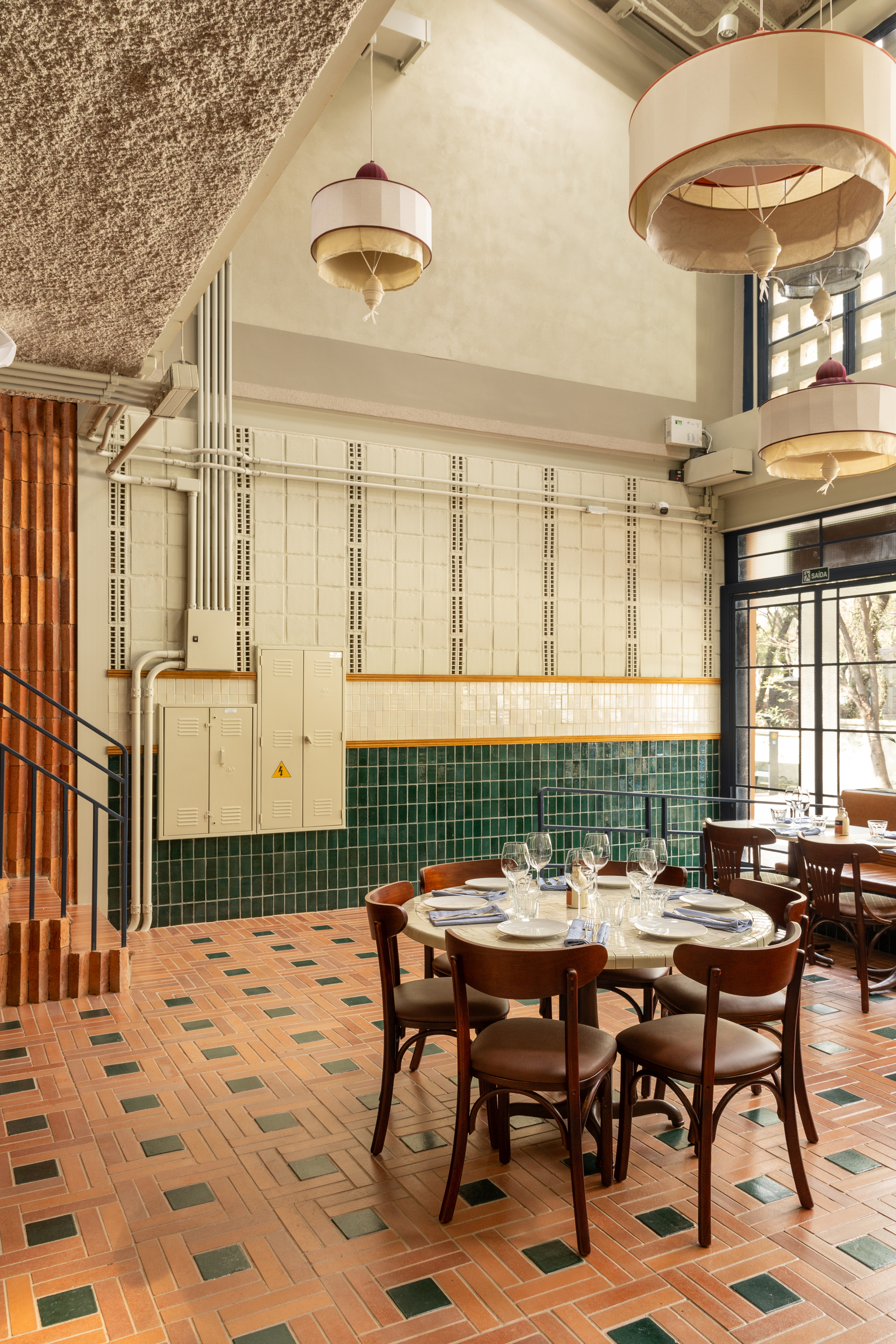 Double-height dining room with green tile wainscot, drum pendant lamps and a round table on terracotta floor tiles, daylight from the tall window