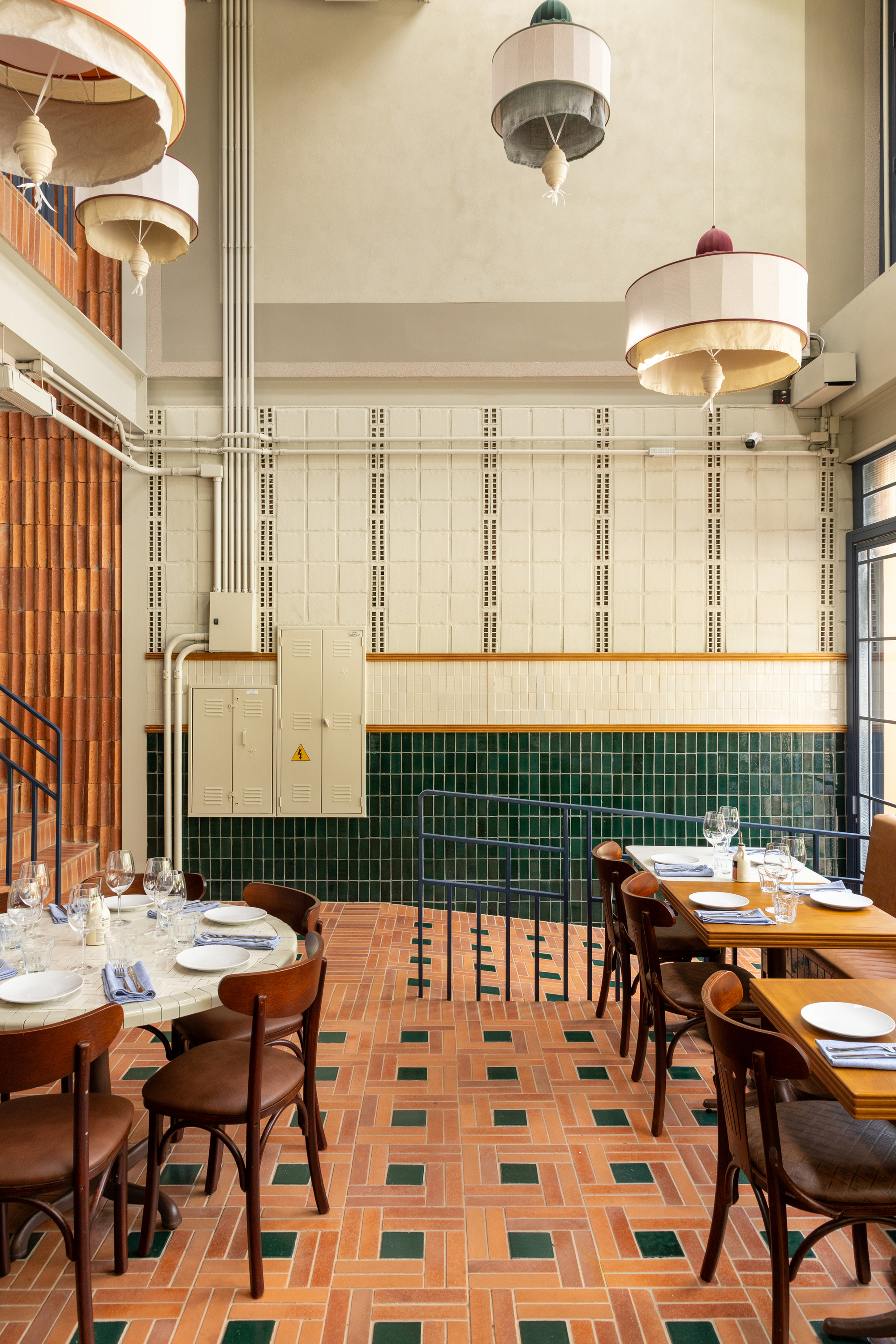 Tall tiled wall with vertical vent strips, an electrical cabinet and drum pendants above tables on a green-inlaid terracotta floor
