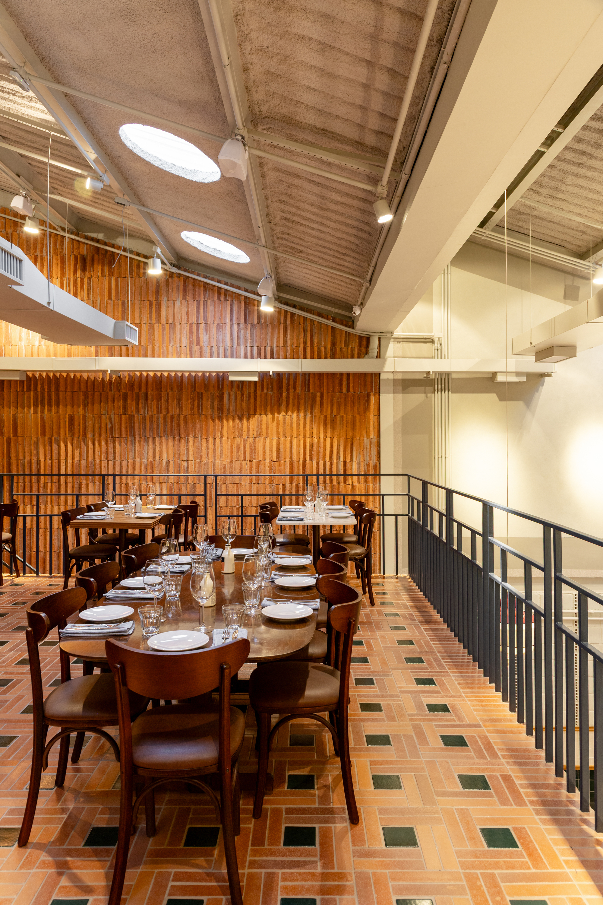Mezzanine dining table beside a dark steel railing, facing a terracotta slat wall under a ceiling with round skylights