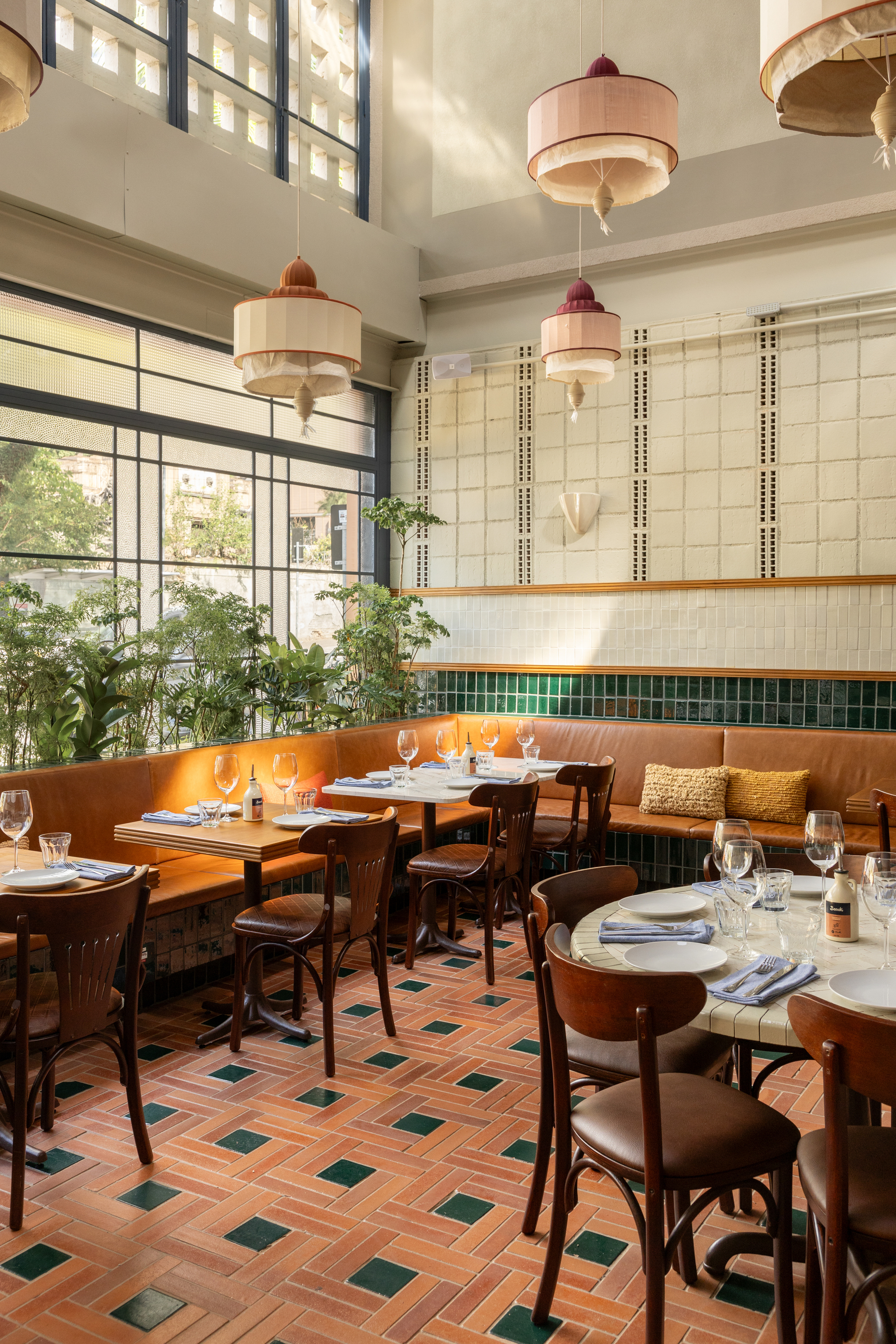 Leather banquette and bentwood chairs along a window with potted plants, under pink and cream pendant lamps in soft daylight