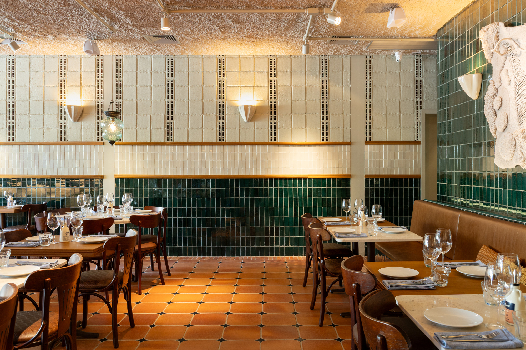 Dining room wall of cream and deep green glazed tiles with brass trim, wall sconces and a relief sculpture in warm evening light