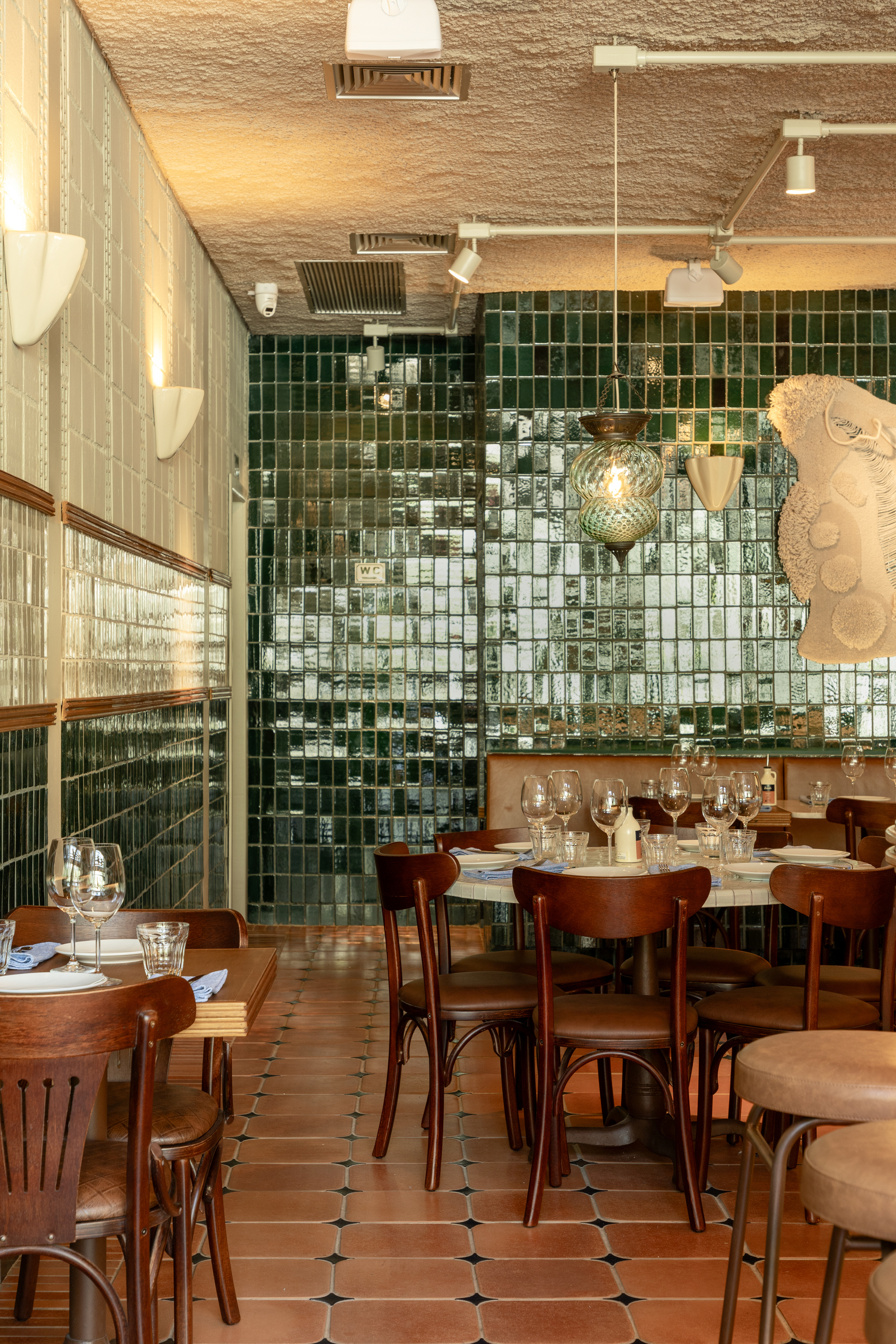 Glossy green tile wall reflecting light behind round tables, with a glass pendant lamp and a pale sculptural relief at right