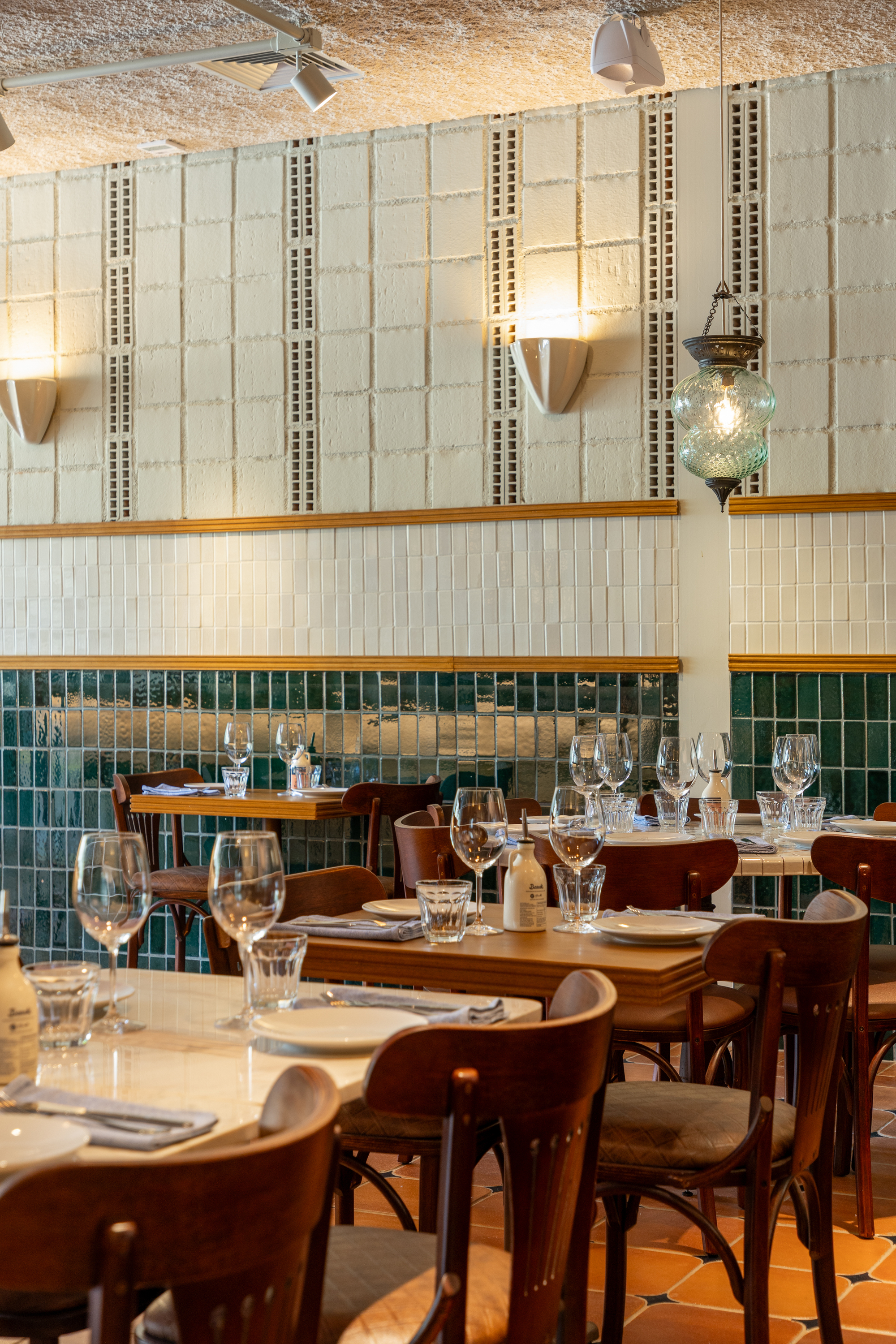 Dining tables and bentwood chairs against a cream tile wall with perforated vertical bands and a green glazed tile dado, lit by wall sconces
