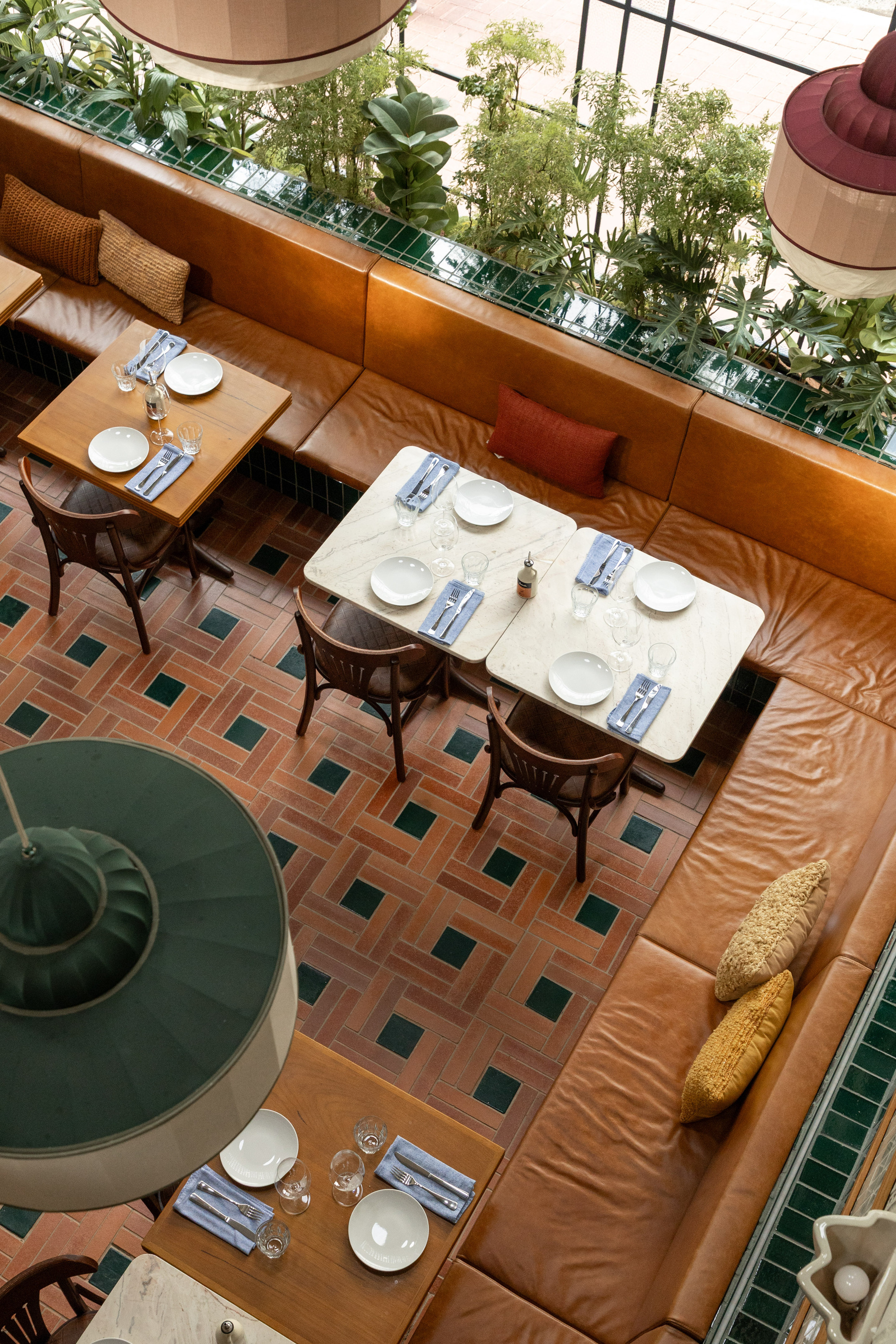 Overhead view of leather banquette seating and marble-topped tables above a herringbone terracotta floor with green inset squares, planting beyond the glass