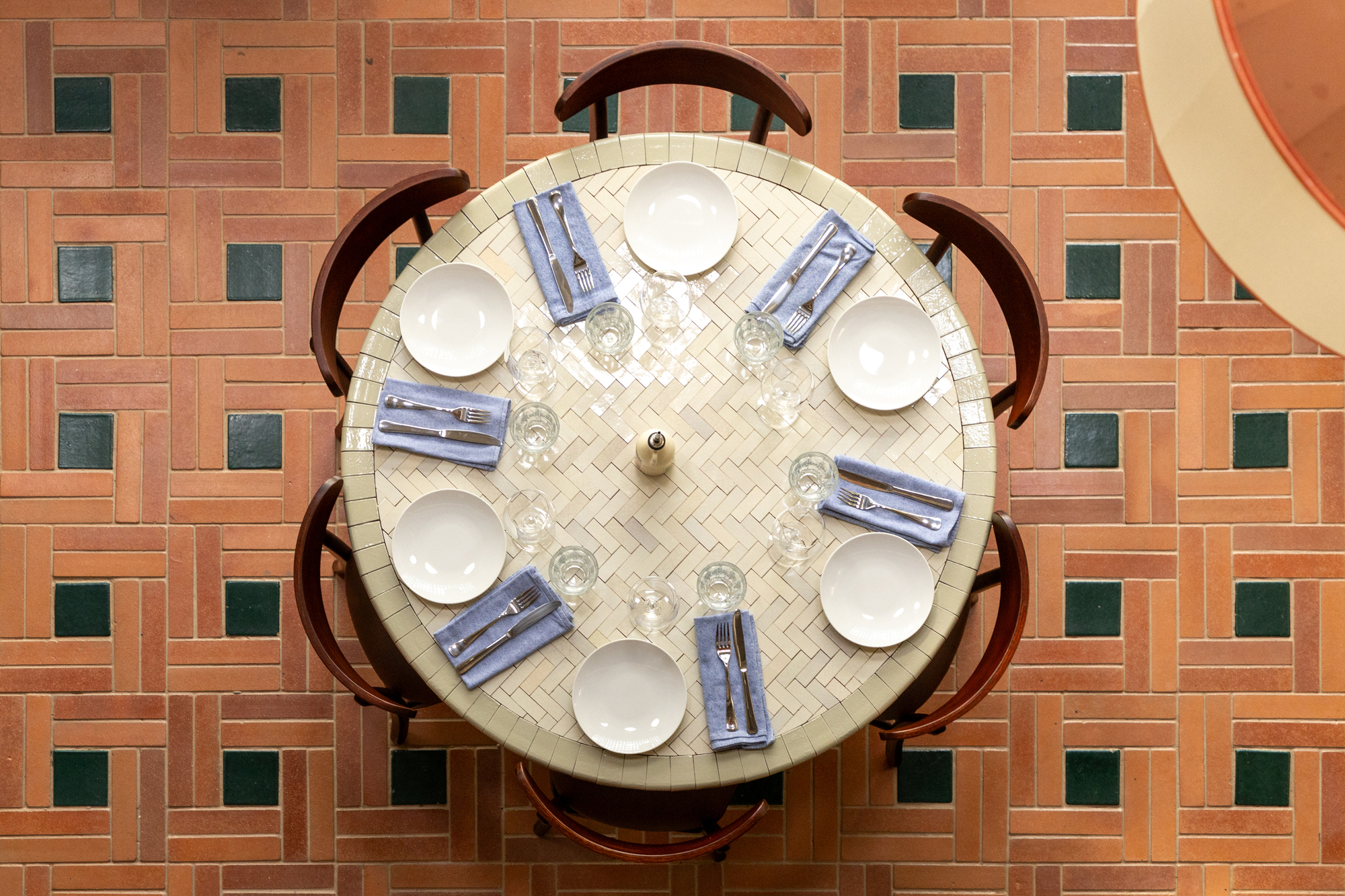 Top-down view of a round tiled table set with plates, folded blue napkins and glasses on a brick-pattern floor