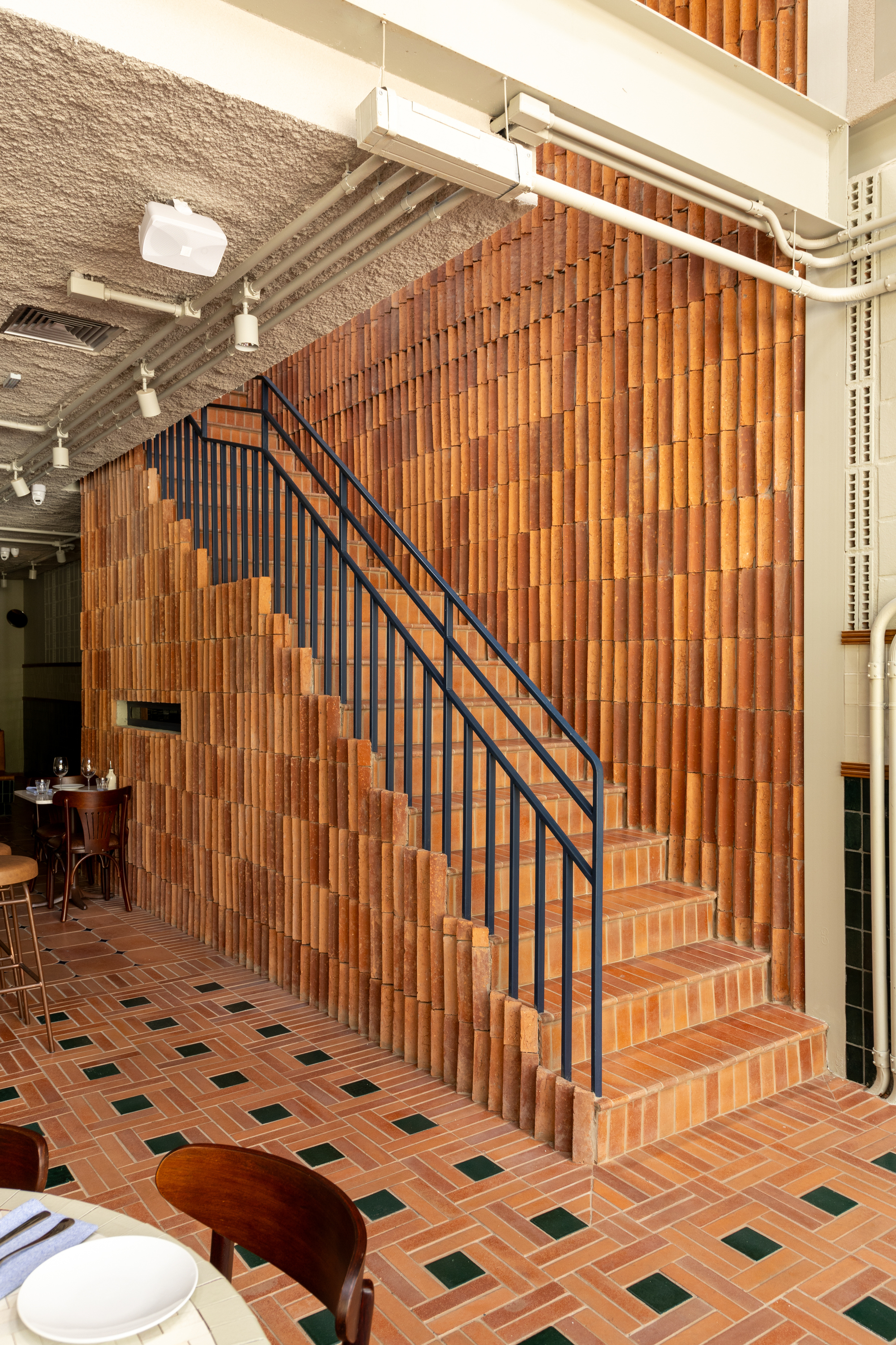 Staircase with dark steel balustrade rising beside a wall of vertically stacked terracotta tiles, exposed pipes running along the ceiling