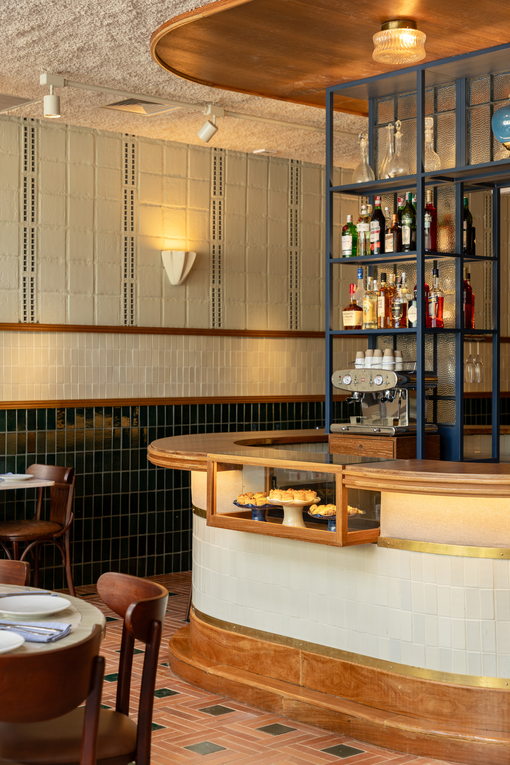 Curved bar with cream tile front and brass trim, a steel shelving frame of bottles above it, and a warm timber canopy