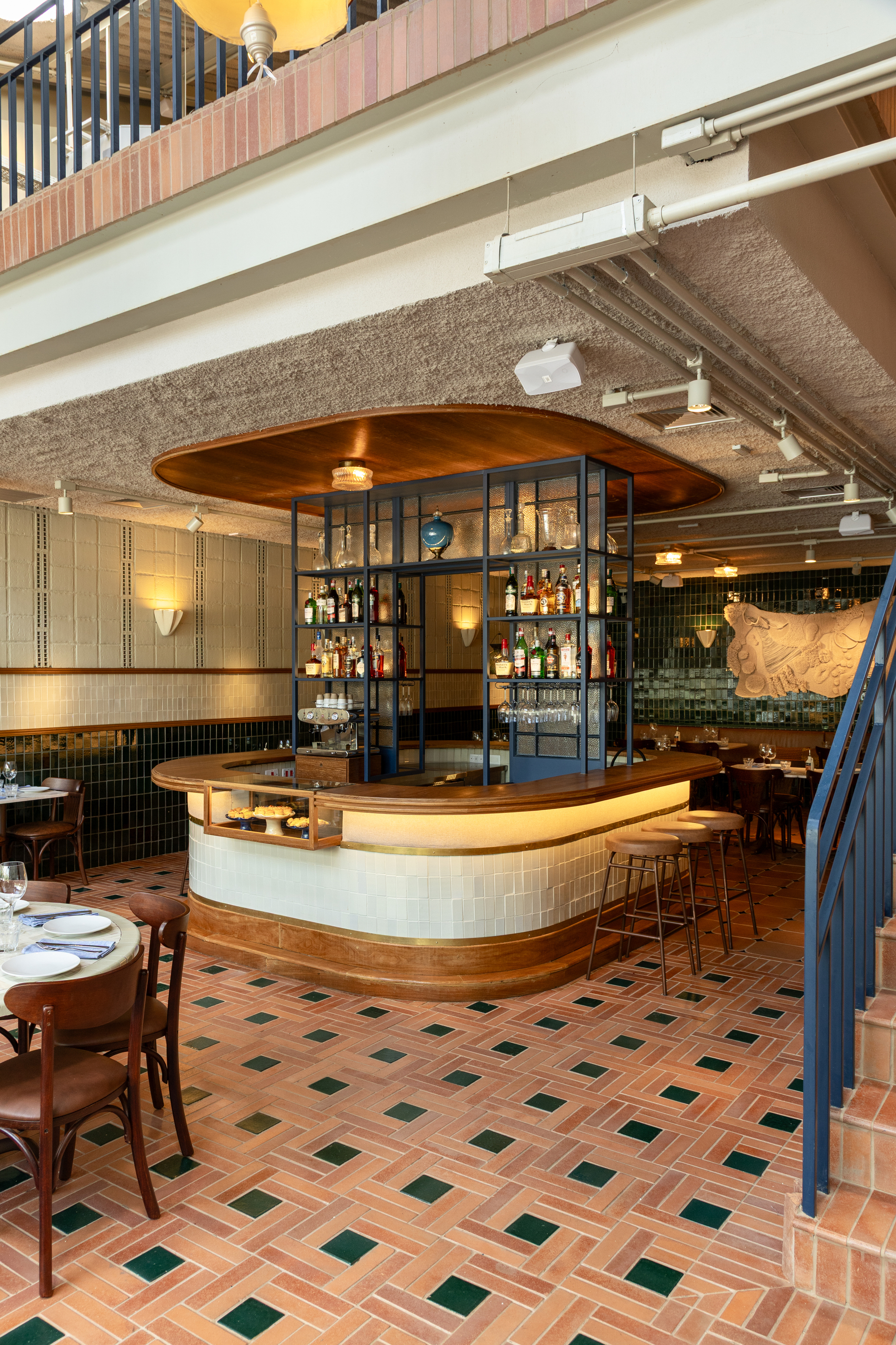 Wide view of the round bar with timber canopy and bar stools, set on a terracotta floor with green squares beneath a mezzanine