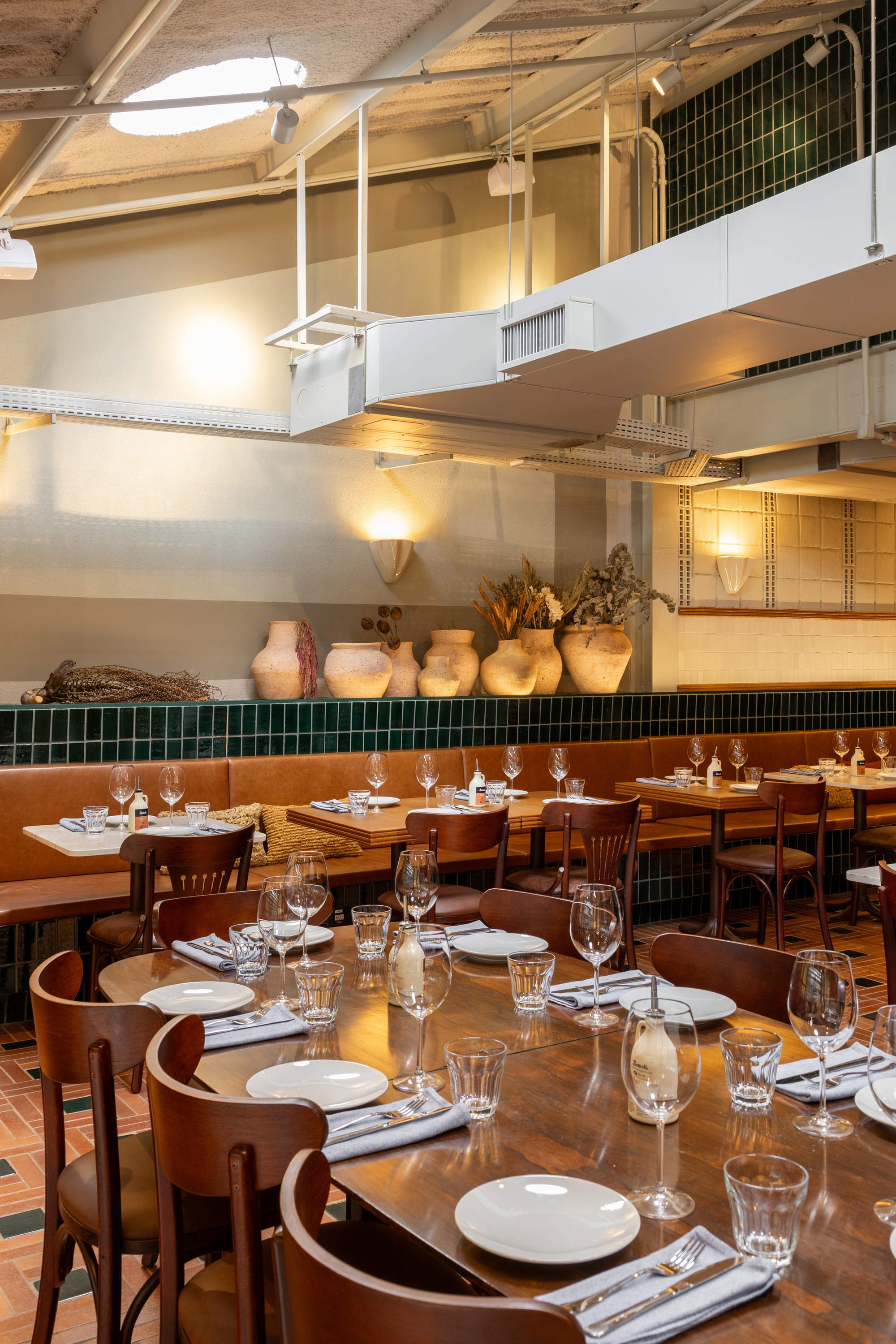 Dining room with wooden tables, bentwood chairs and a leather banquette against a green tile dado, clay vases glowing under wall sconces