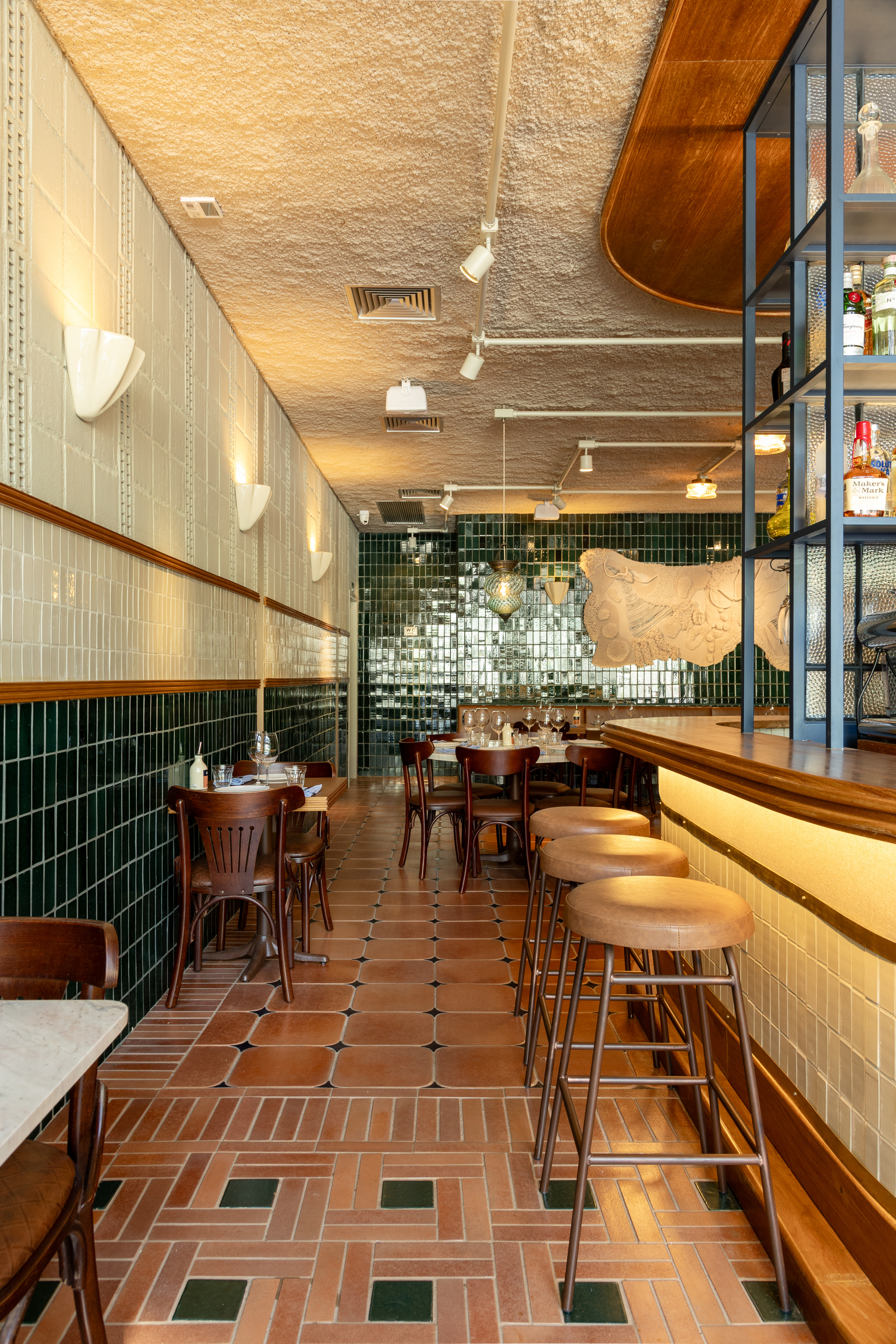 Long restaurant interior with terracotta herringbone floor, green tile wall base, leather bar stools and a curved timber bar edge under warm track lights