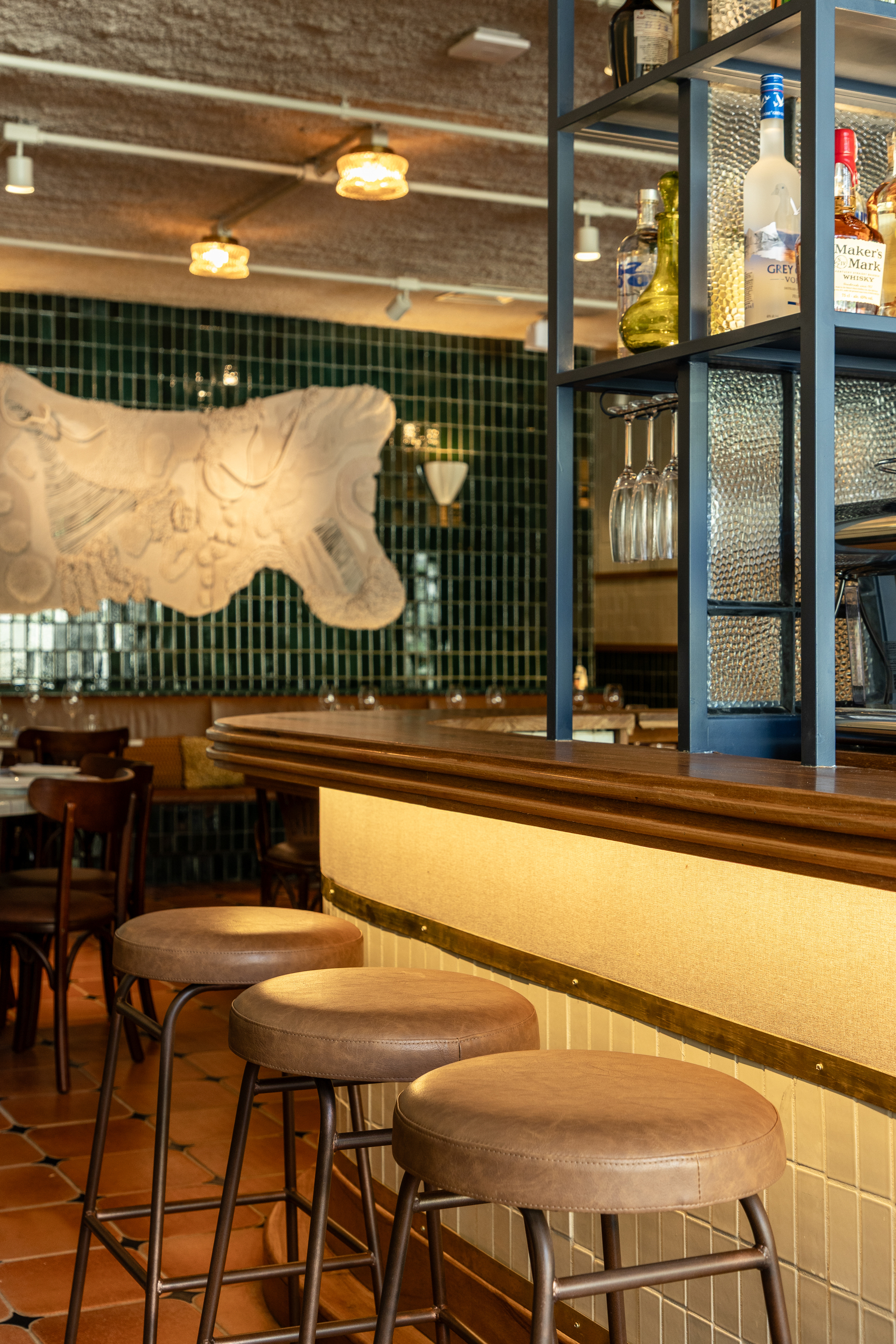 Three leather-topped bar stools at a curved bar with a brass trim, a bottle shelf and a relief panel on green tile behind