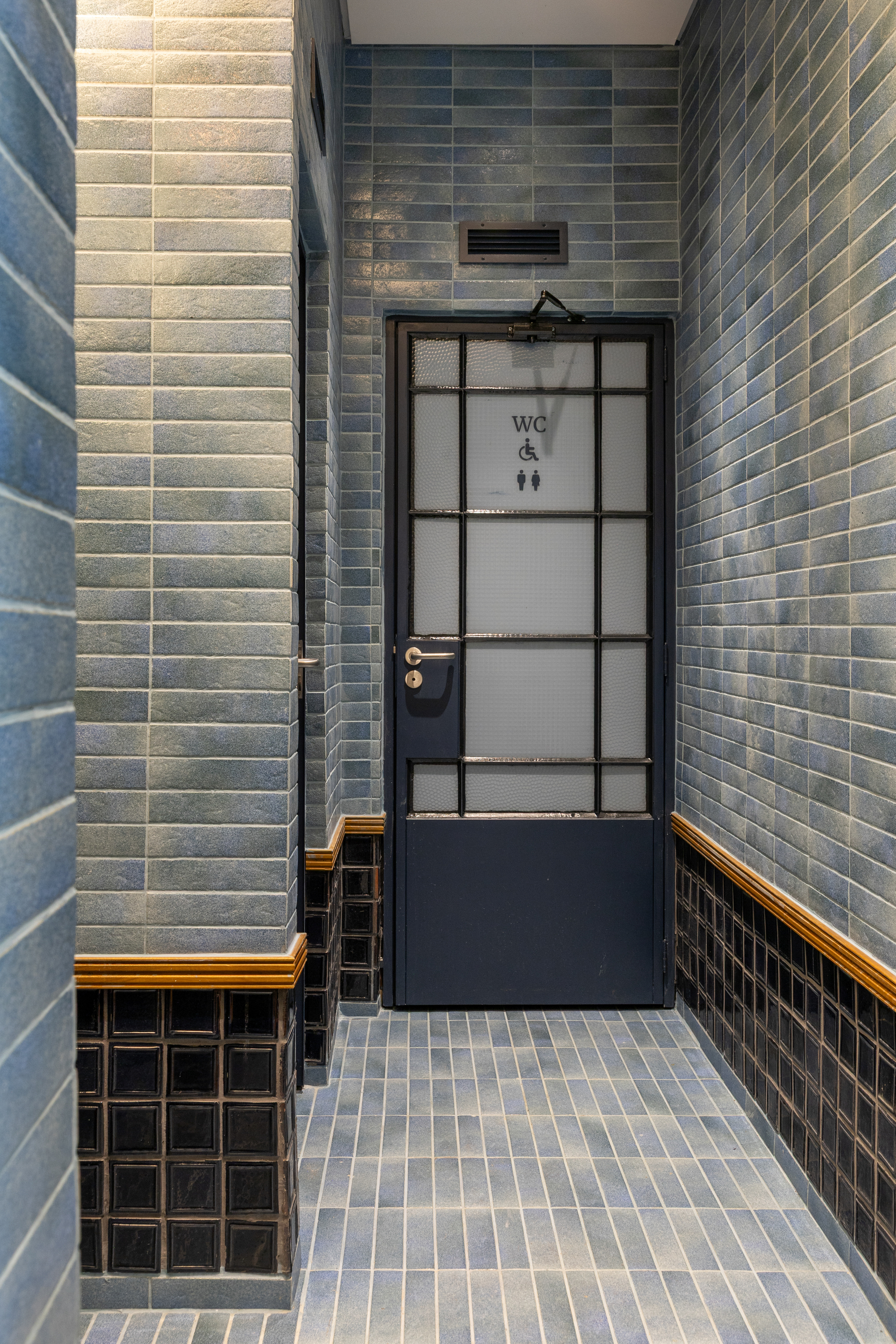 Restroom corridor clad in blue glazed tile with a dark steel-framed frosted glass door marked WC and a timber trim band