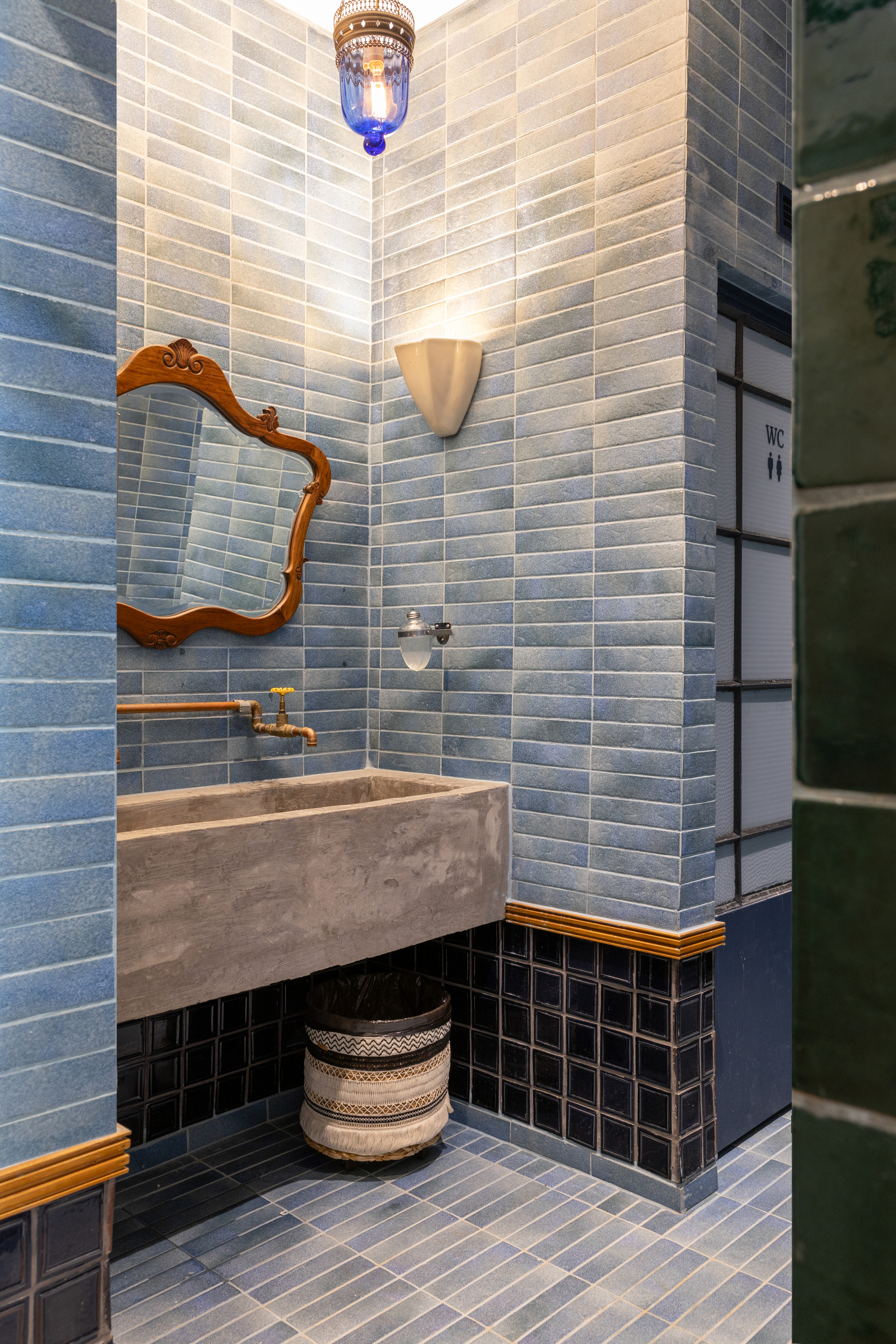 Washroom corner with a concrete trough sink, ornate wooden mirror and a blue glass pendant lamp over blue tiled walls