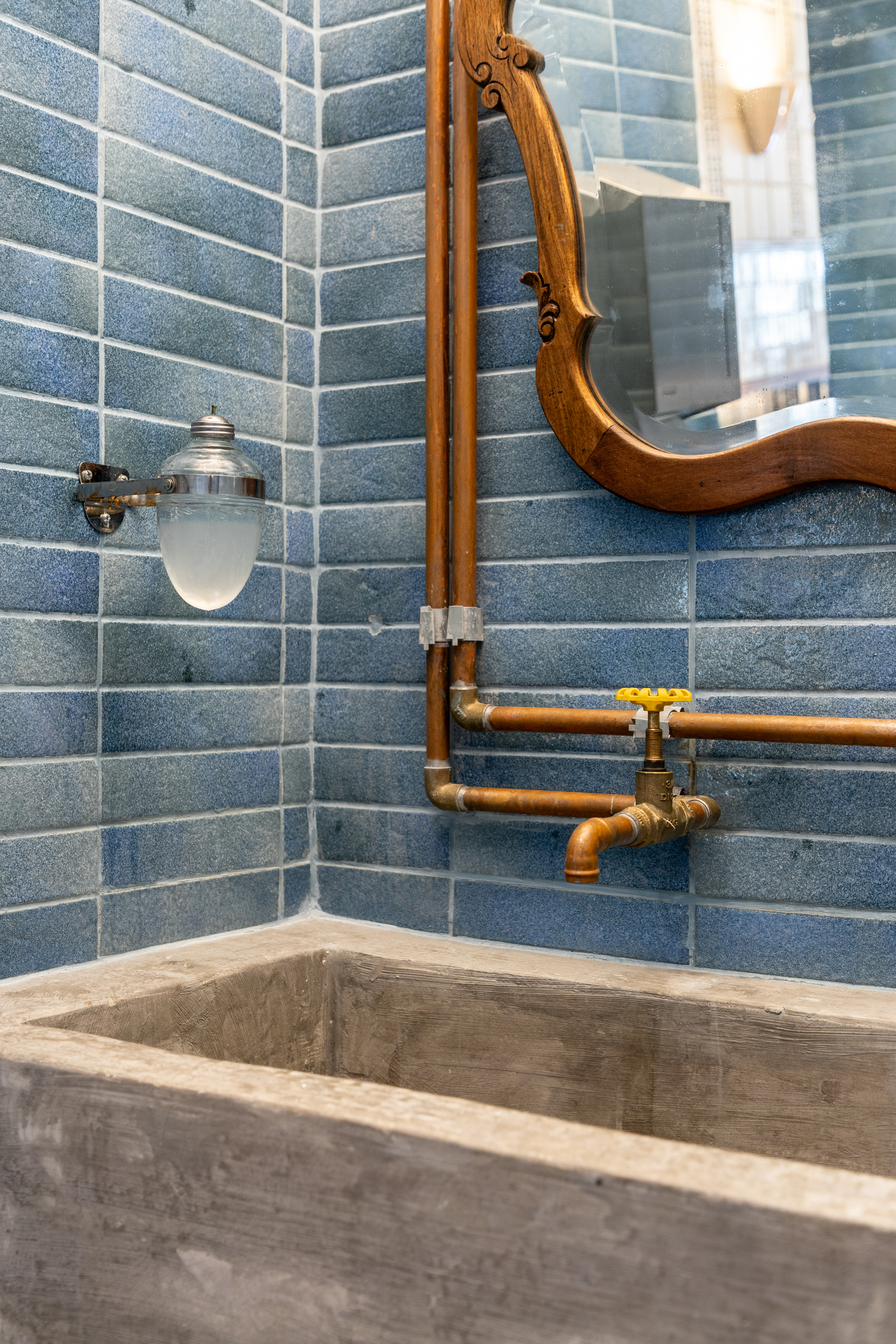 Close-up of exposed copper pipes and a yellow-handled brass tap above a concrete trough sink, framed by a carved wooden mirror
