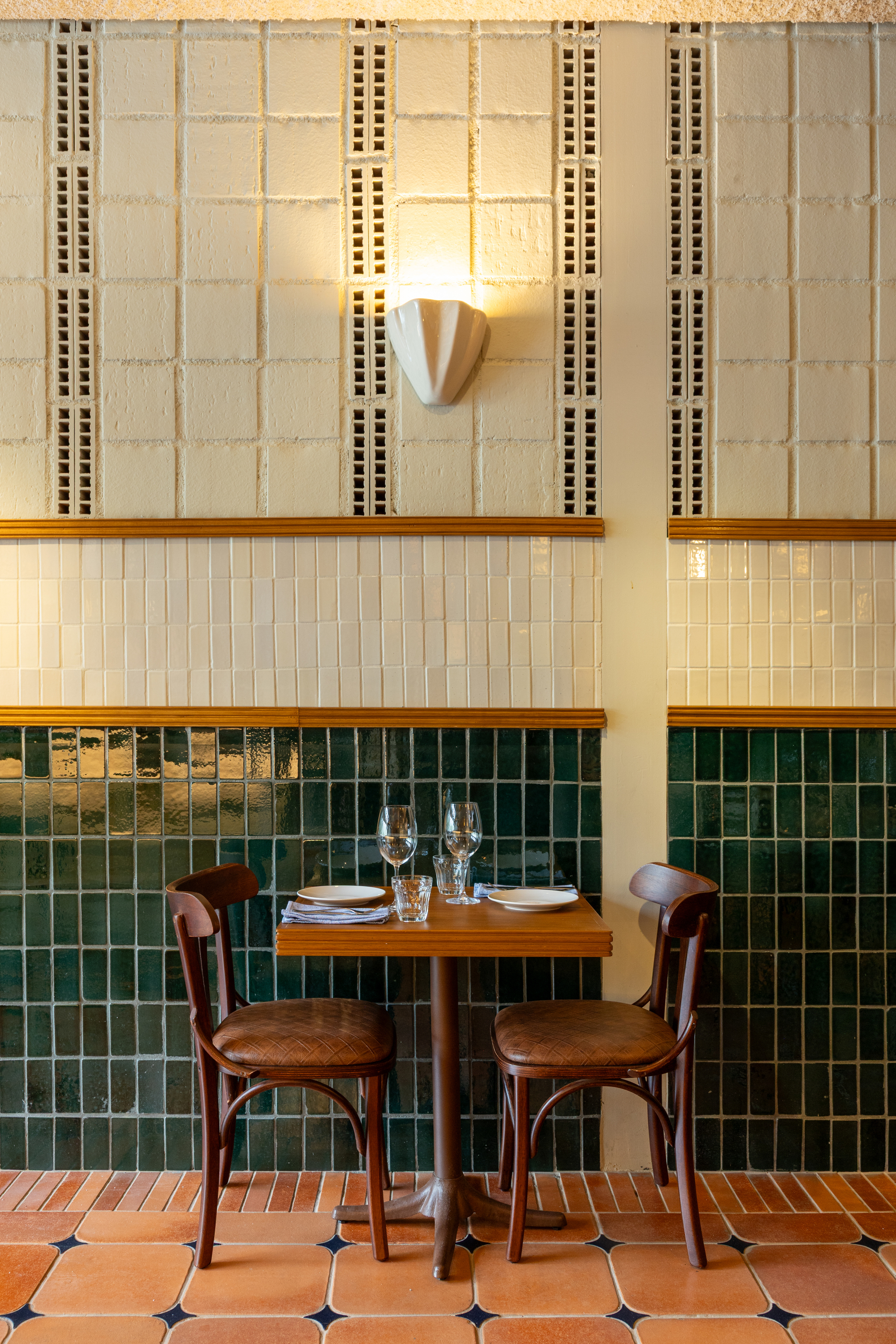 Two bentwood chairs at a small timber table against a wall of green and cream glazed tiles under a shell-shaped sconce