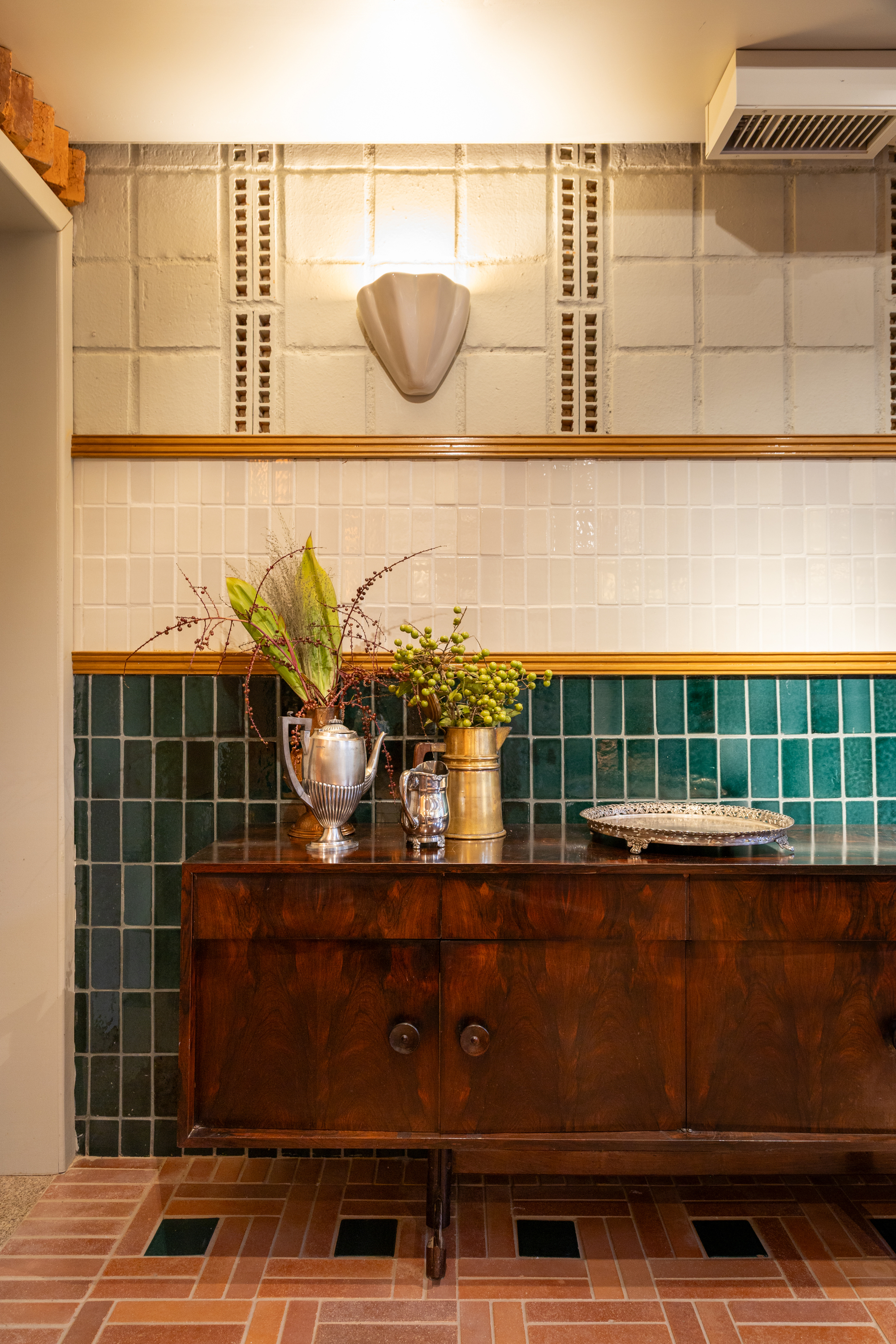 Walnut sideboard holding silver pots and dried foliage in front of green tile, with a terracotta floor inset with black squares