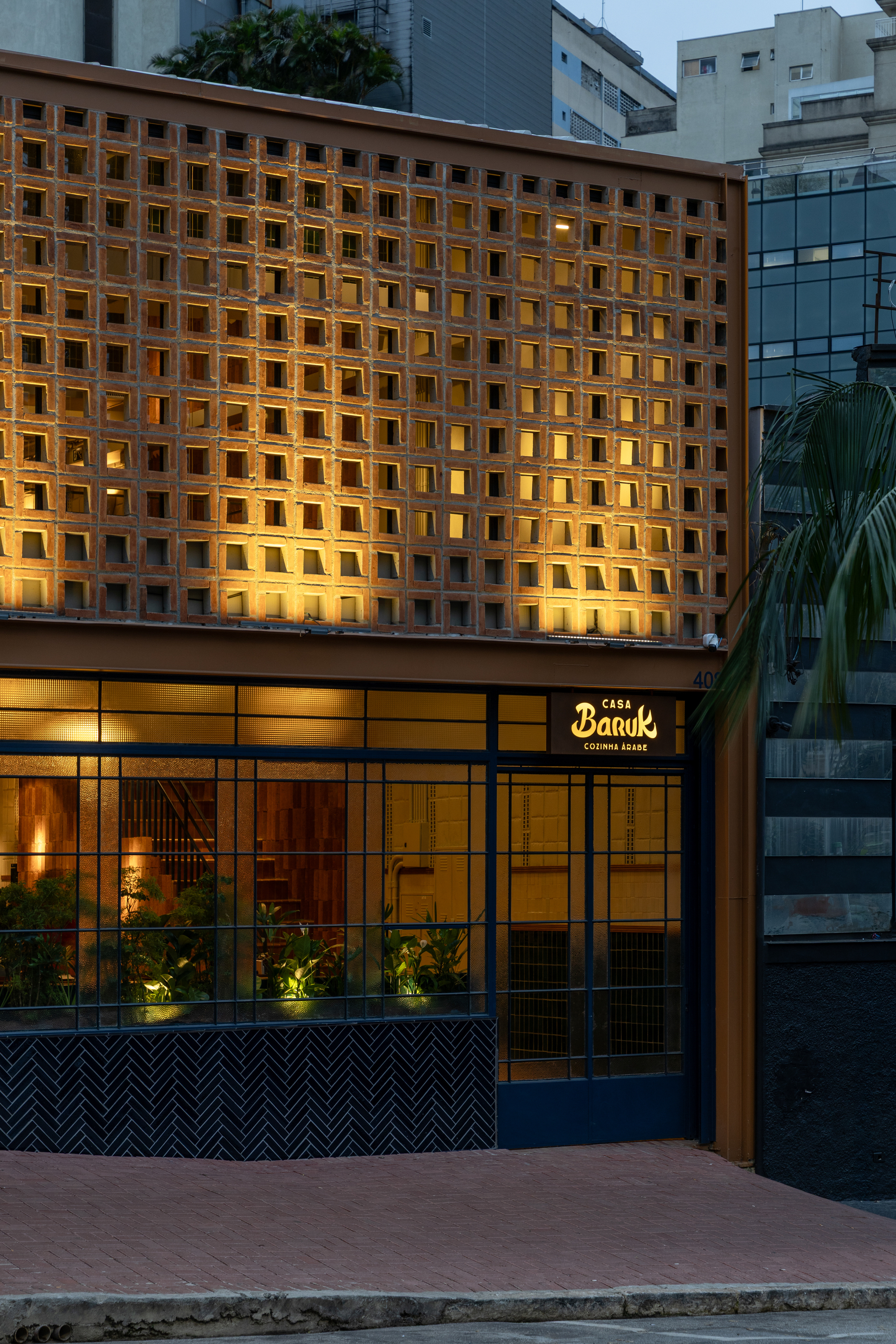 Street facade at dusk with a lit perforated block screen above a blue steel-framed glass front and planted interior