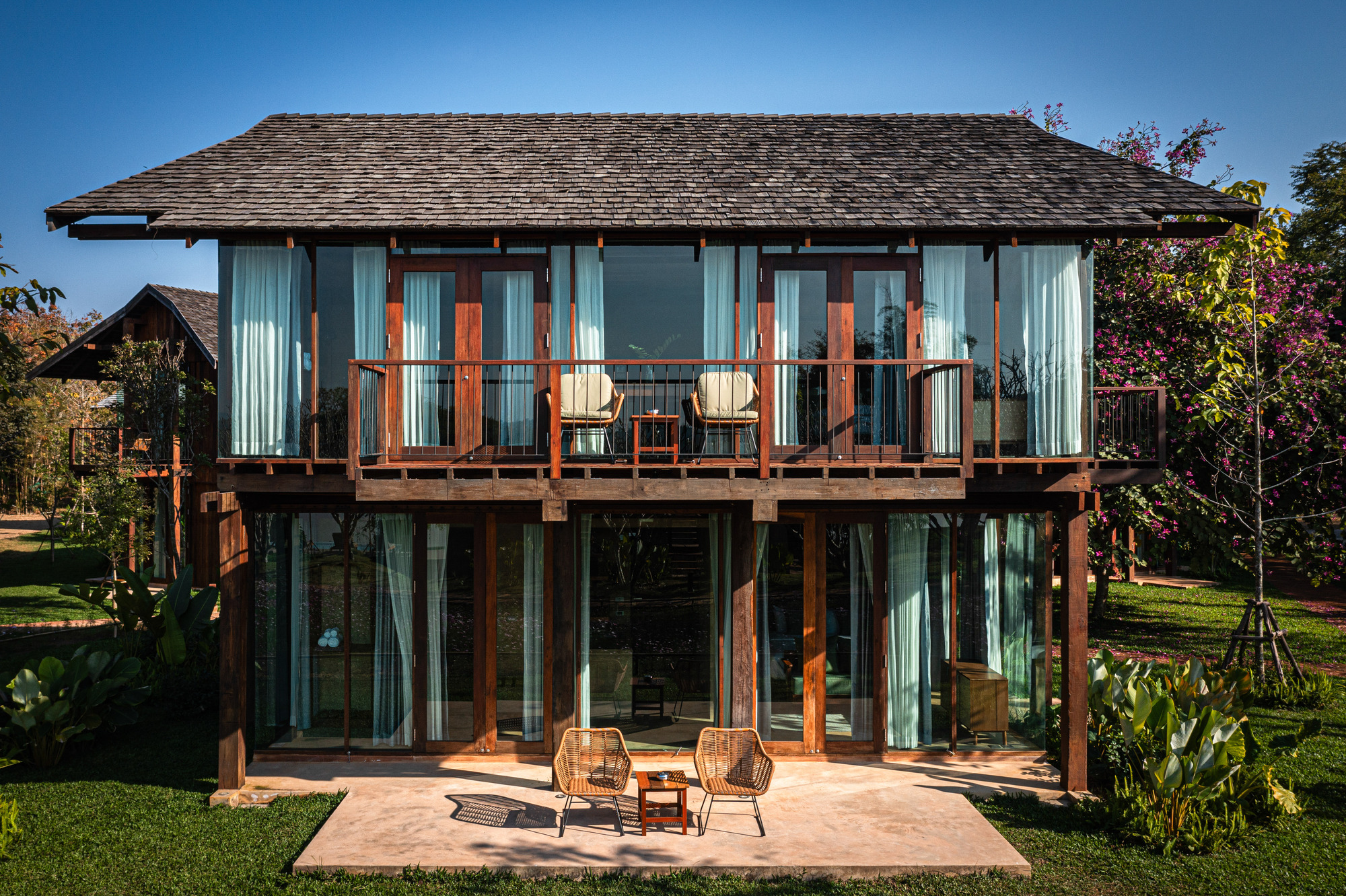 Two-storey timber pavilion with shingle roof, upper balcony and teal curtains, fronted by a concrete terrace and two rattan chairs under clear blue sky