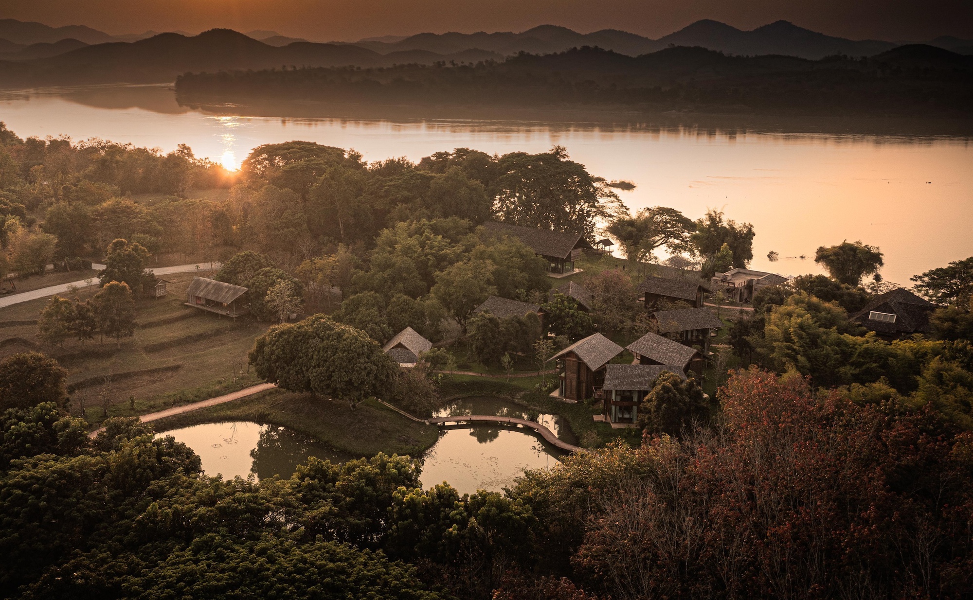 Aerial view of timber cabins with pitched roofs among trees beside a pond, with the sun setting over a wide river